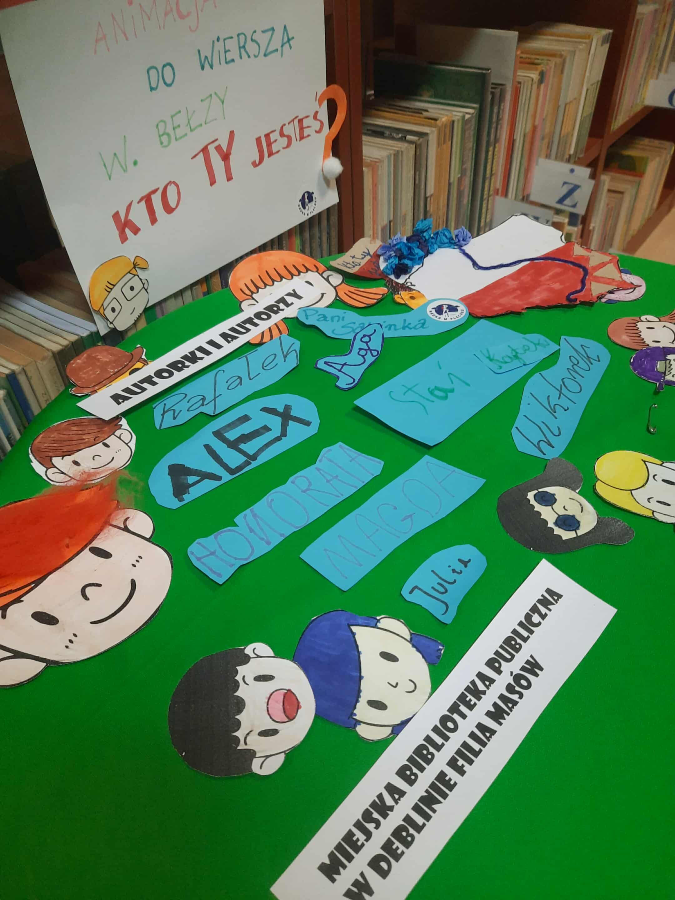 Kolorowe, ręcznie wykonane ozdoby, napisy i postacie z papieru leżą na zielonym tle. W tle widać półki z książkami. Na przyczepionej kartce napis: "ANIMACJA DO WIERSZA... KTO TY JESTEŚ?" oraz informacje o bibliotece.