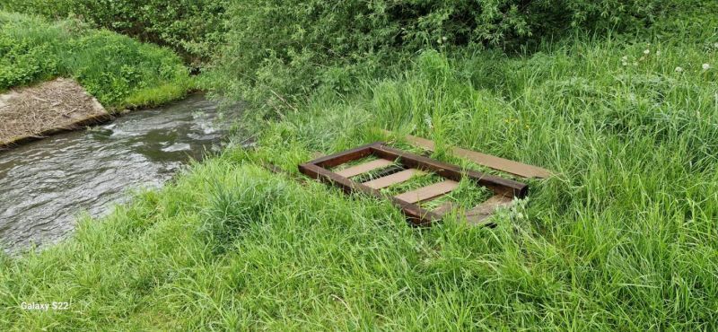 Zdjęcie przedstawia drewnianą kładkę nad strumieniem otoczoną bujną, zieloną trawą i roślinnością.