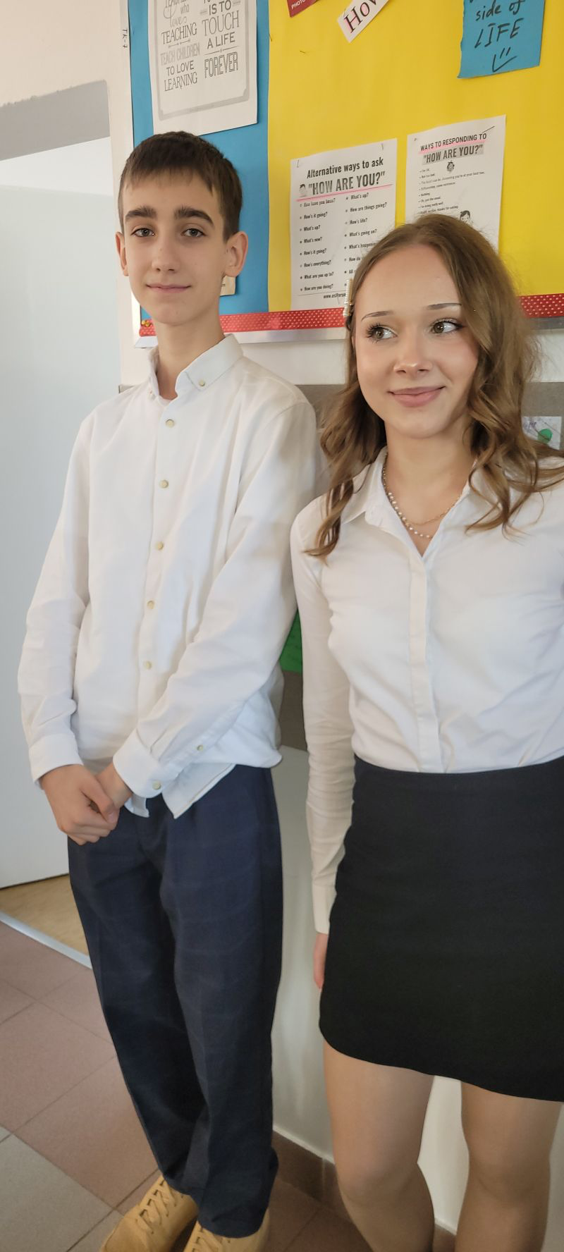 Dwóch młodych ludzi w eleganckich ubraniach stoi obok siebie; chłopak w białej koszuli i granatowych spodniach, dziewczyna w białej koszuli i czarnej spódnicy.