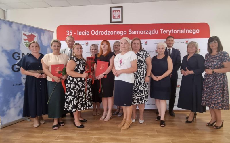 Grupa osób pozuje razem na tle banera z napisem "35-lecie Odrodzonego Samorządu Terytorialnego". Niektórzy trzymają czerwone kwiaty i teczki z dokumentami. W tle znajduje się flaga z godłem gminy.