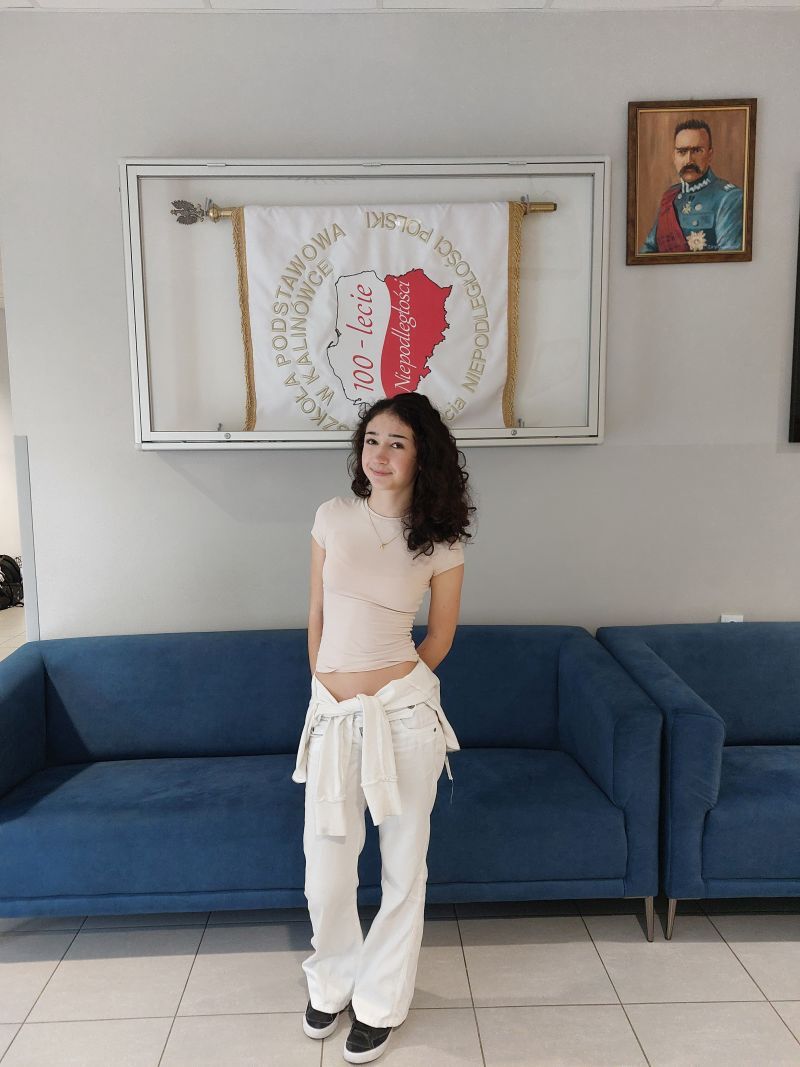 Młoda kobieta z ciemnymi włosami stoi przed niebieską kanapą. Ma na sobie jasną bluzkę i białe spodnie. W tle widoczna jest tablica z napisem, a na ścianie wisi portret mężczyzny w mundurze.