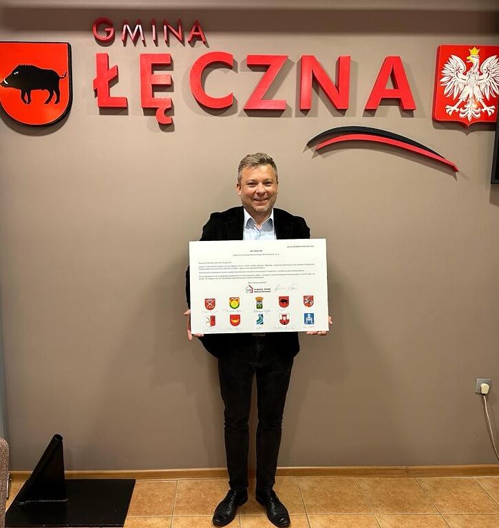 Mężczyzna uśmiecha się i trzyma duży czek z herbami. W tle czerwona ściana z napisem "Gmina Łęczna" i herbem Polski.