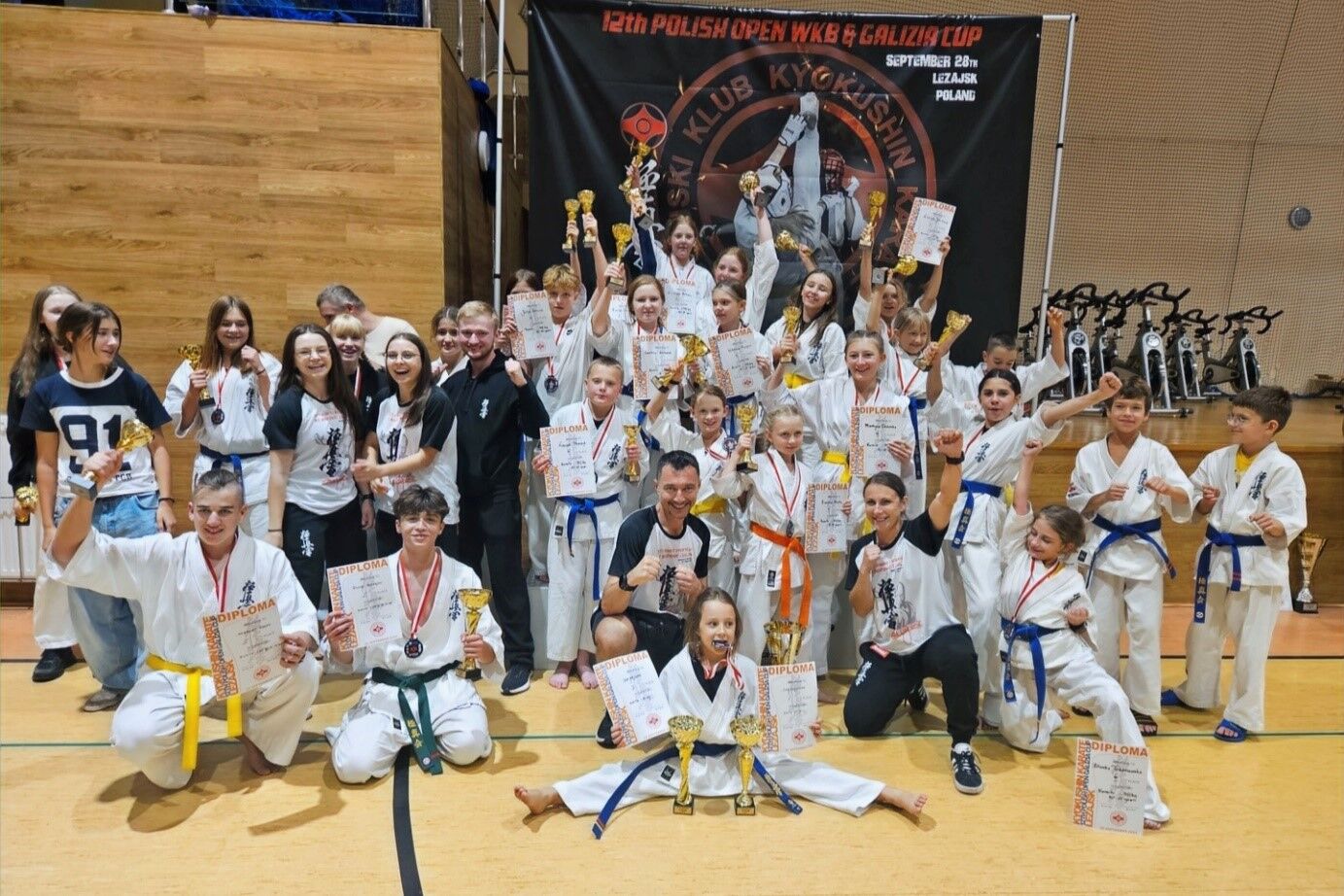 Grupa osób w kimono karate stoi na sali gimnastycznej, trzymając trofea i medale, z banerem zawodów w tle.