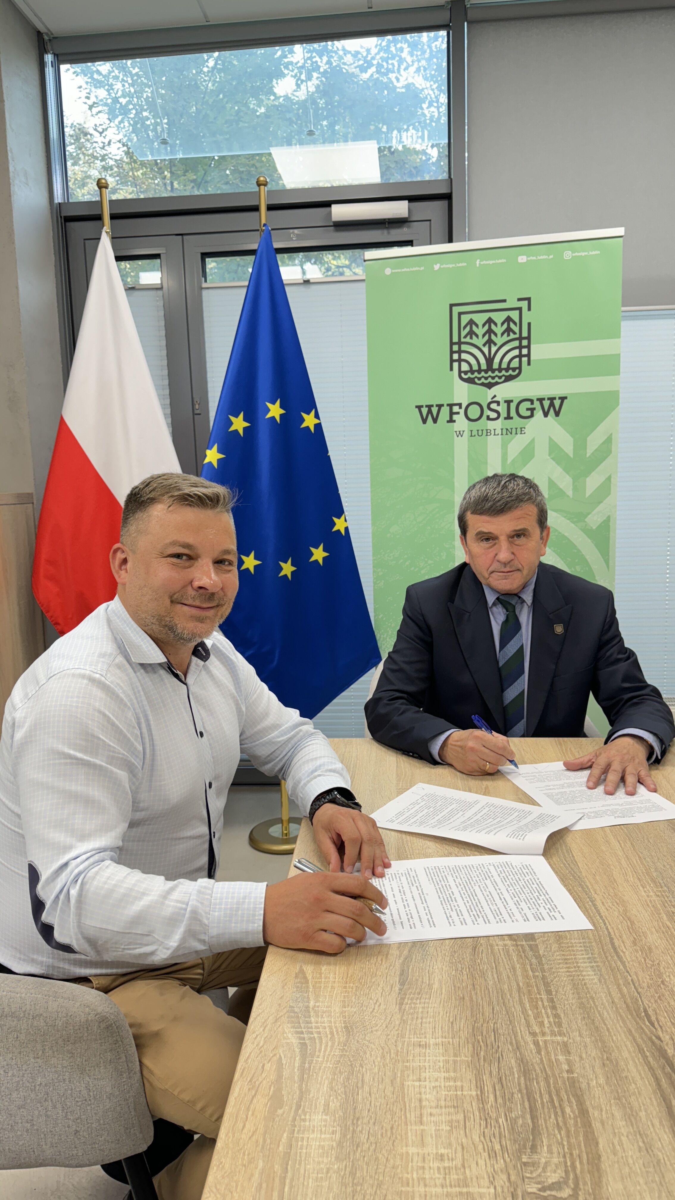 Dwaj mężczyźni siedzą przy stole w biurowym pomieszczeniu, jeden podpisuje dokument. Polska i UE flagi w tle, a także logo WFOSiGW.