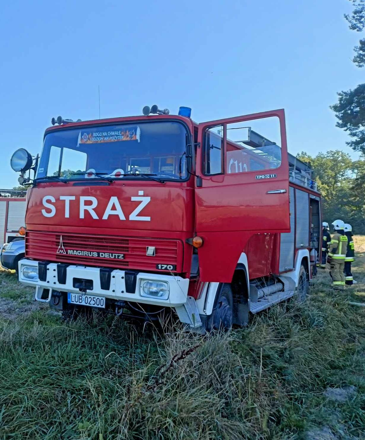 Czerwony wóz strażacki Magirus Deutz stojący na trawiastym terenie z widocznymi strażakami w tle.