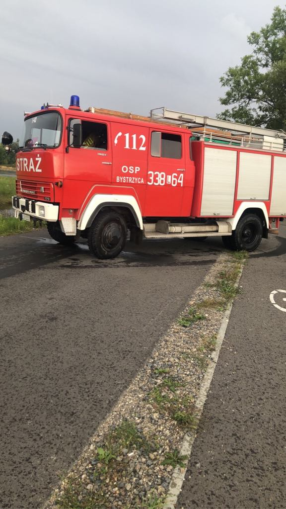 Czerwony wóz strażacki OSP z numerem 338*64 zaparkowany na asfaltowej drodze, z napisem "112 STRAŻ" oraz oznaczeniem miejscowości "BYSTRZYCA".