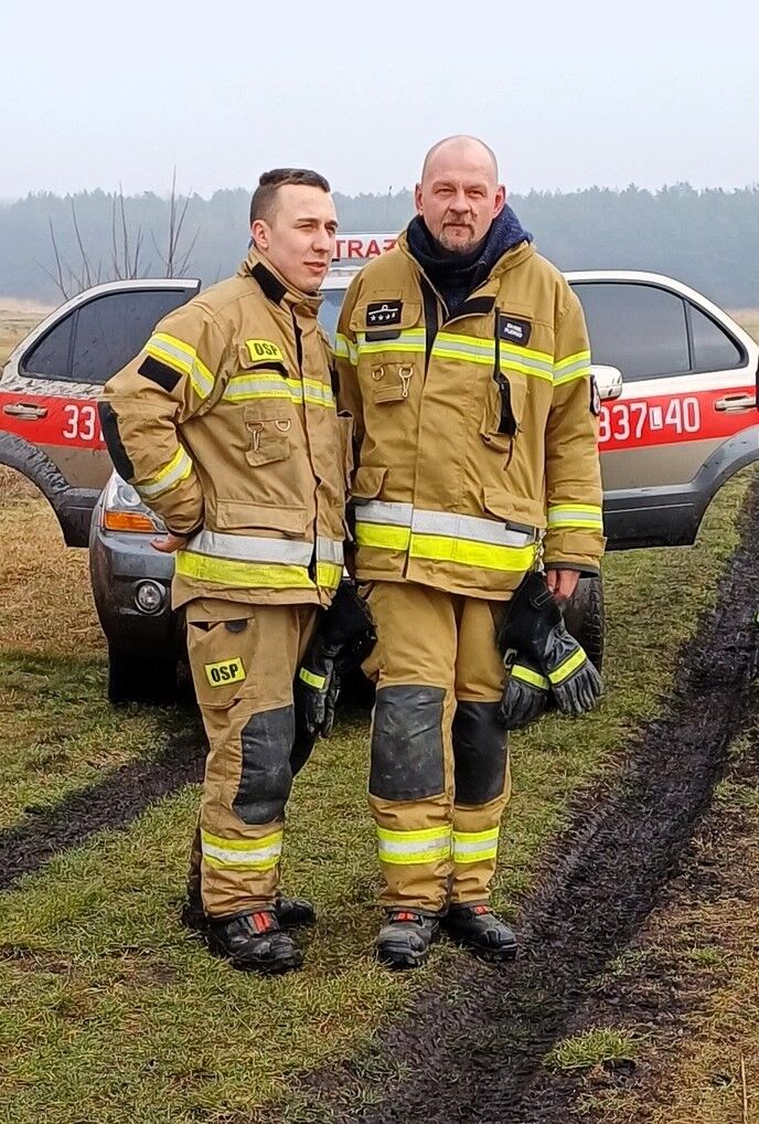 Dwóch strażaków w pełnym wyposażeniu stoi na trawiastym terenie z samochodem pożarniczym w tle.
