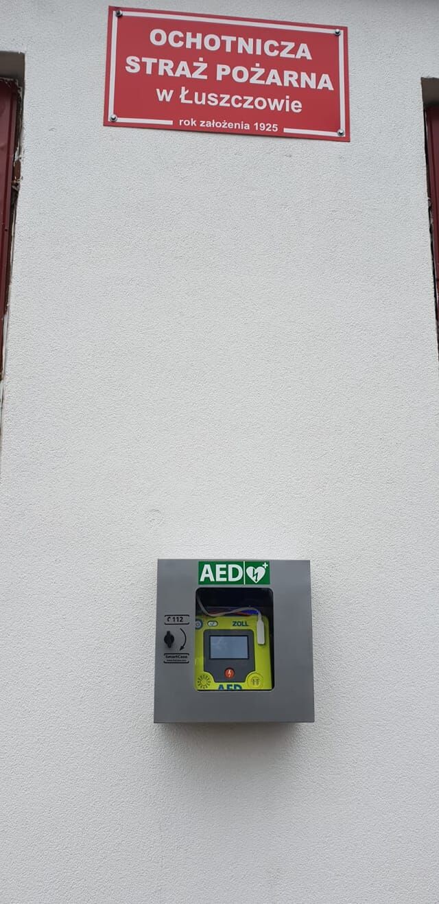 Defibrylator AED zamontowany na ścianie zewnętrznej, pod znakiem "Ochotnicza Straż Pożarna w Łuszczowie", data założenia 1925.