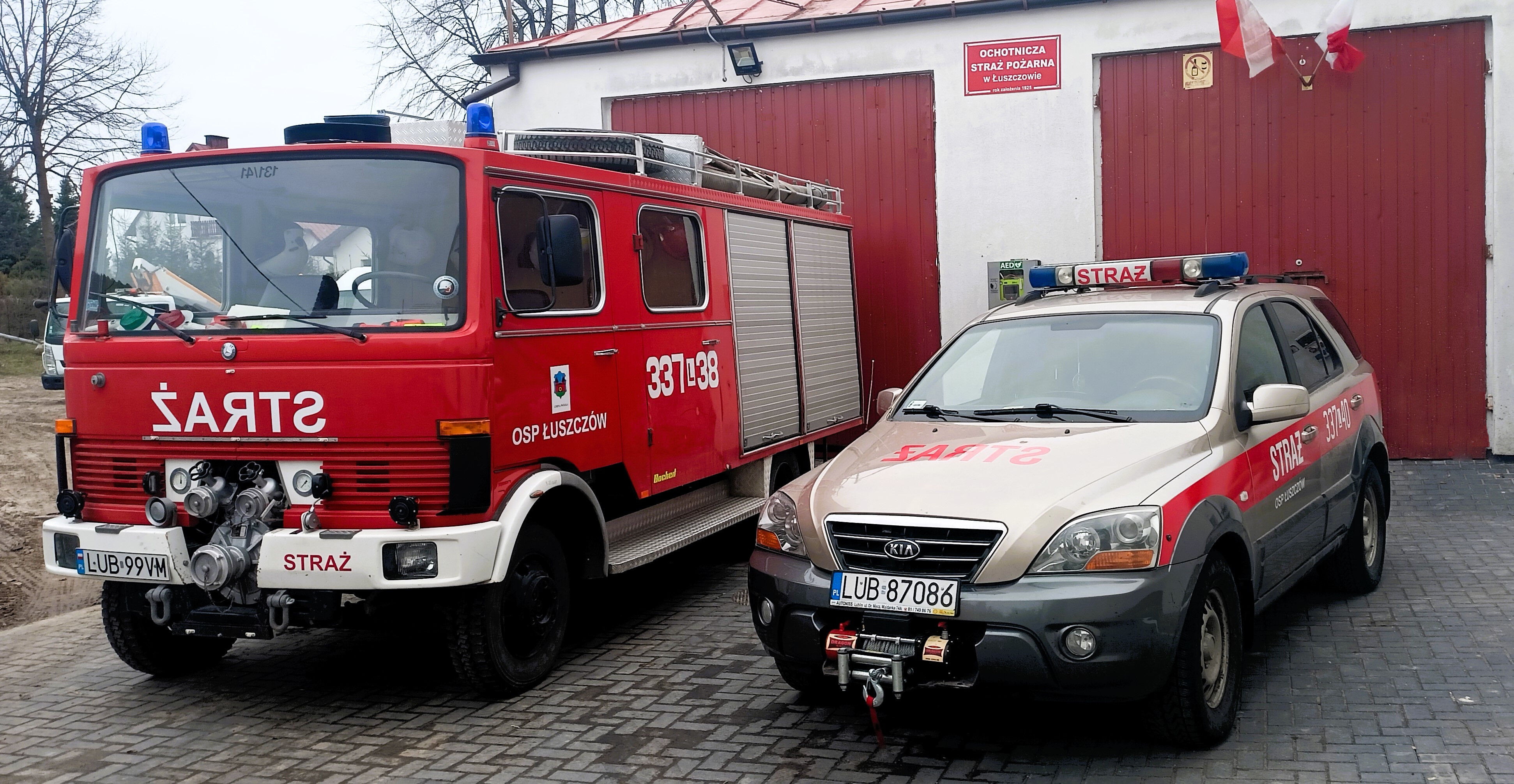 Czerwony wóz strażacki i srebrny samochód operacyjny OSP z napisem "Straż" przed budynkiem straży pożarnej.