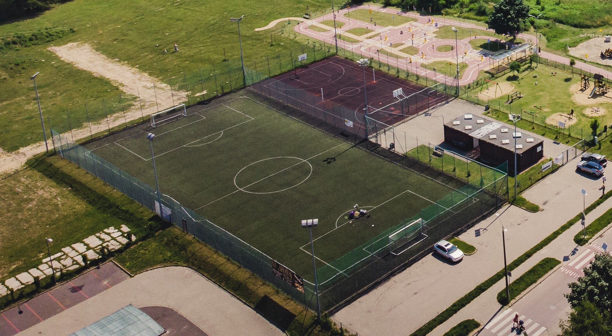 Zdjęcie lotnicze z boiskiem do piłki nożnej z sztuczną nawierzchnią, boiskami do koszykówki, placem zabaw oraz parkiem i parkingiem.