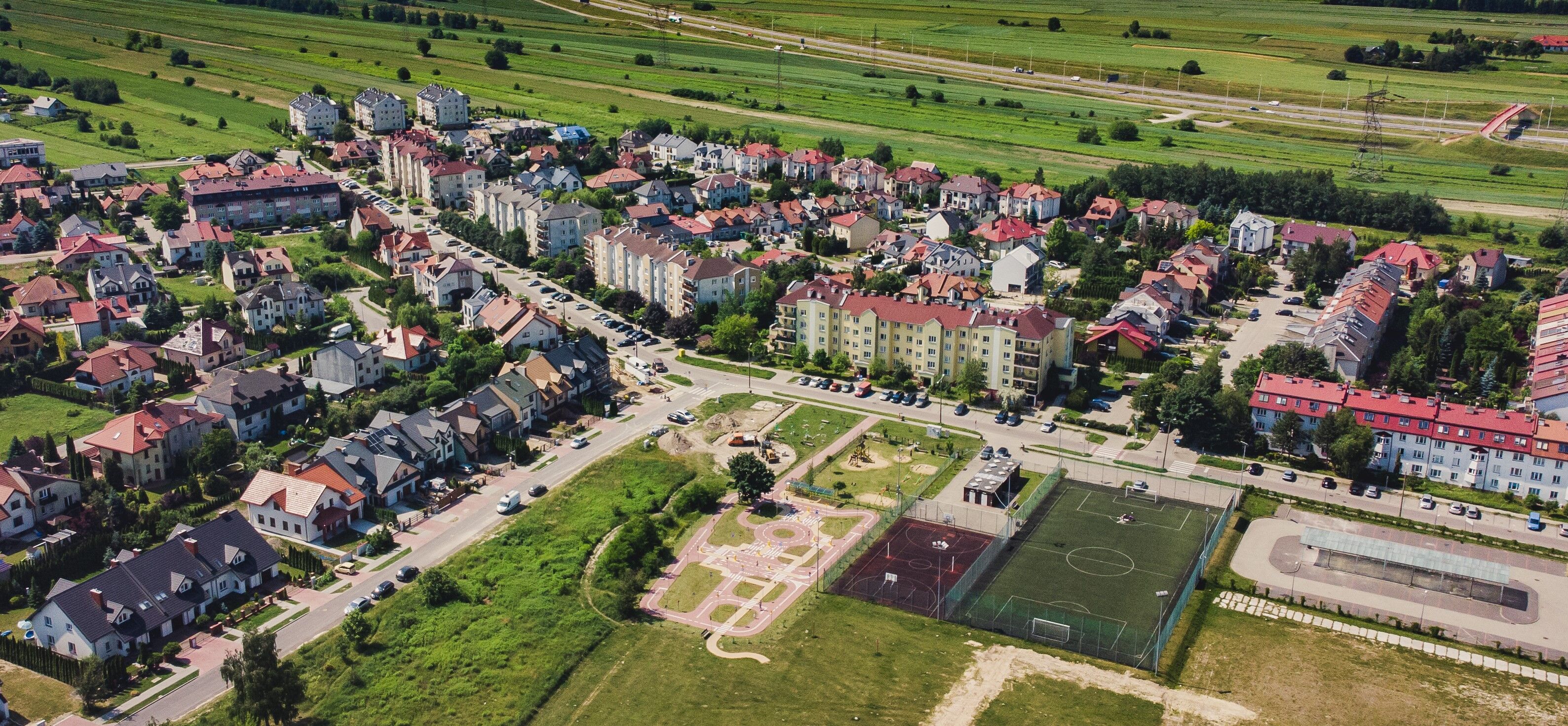 Widok z lotu ptaka na przedmieścia: mieszane budownictwo, boisko sportowe, zieleń i ulice.