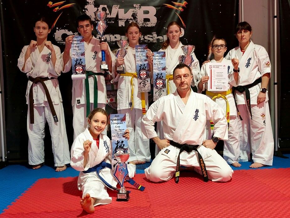 Na zdjęciu grupa osób w strojach do sztuk walki pozuje z pucharami i dyplomami na zawodach. Na pierwszym planie dziewczynka z pucharem, obok niej siedzi mężczyzna. W tle widoczny jest baner z logo turnieju.