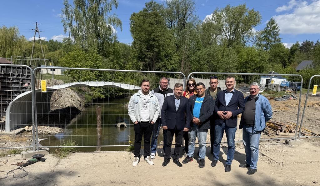 Grupa osób stoi przed ogrodzeniem, za którym znajduje się plac budowy z konstrukcją mostu. Wokół są drzewa, a niebo jest bezchmurne.