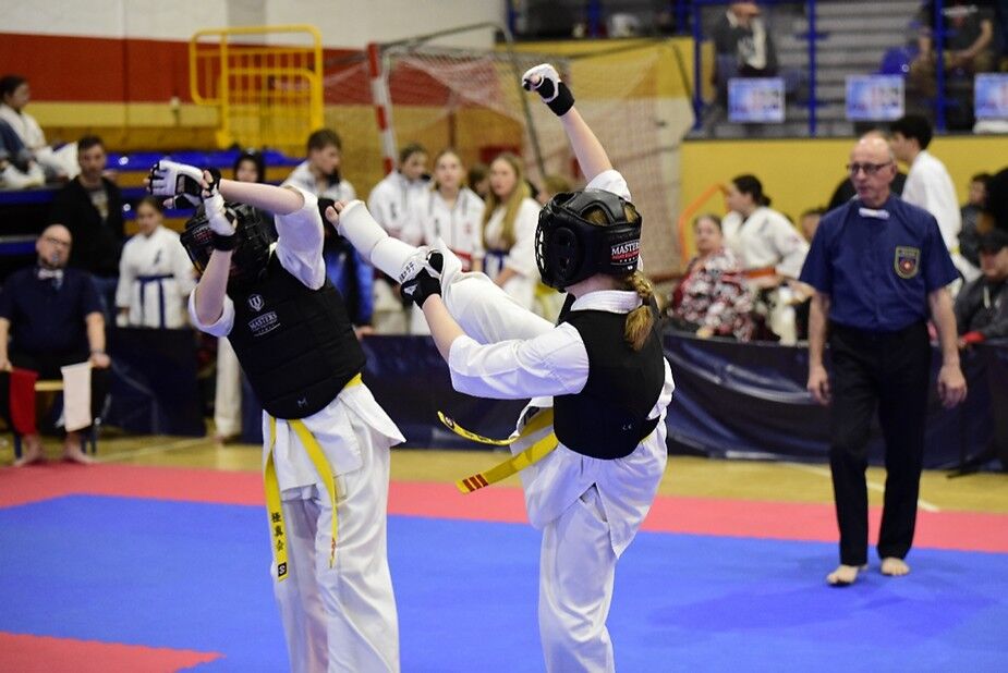 Dwóch zawodników w białych strojach i żółtych pasach walczy na zawodach karate. Jeden z nich wykonuje kopnięcie, drugi unosi ręce w obronie. Na drugim planie widoczni sędziowie i publiczność.