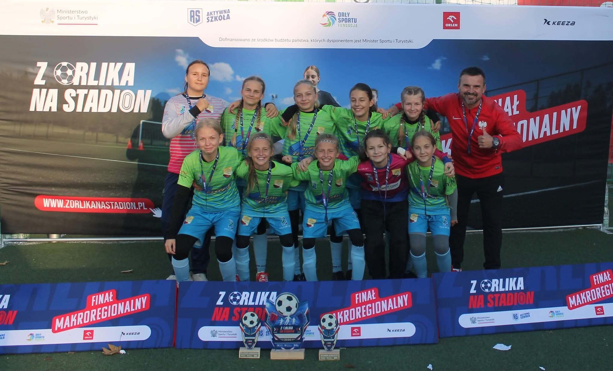 Drużyna dziewcząt w zielono-niebieskich strojach piłkarskich z medalami i trener w czerwonej bluzie przed ścianką Z Orlika na Stadion, Finał makroregionalny. Na pierwszym planie trzy puchary z piłkami.