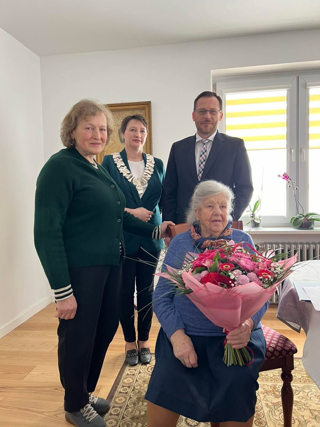 Zdjęcie przedstawia cztery osoby w jasnym pokoju. Na pierwszym planie, na krześle po prawej, siedzi starsza kobieta z siwymi włosami spiętymi z tyłu, ubrana w niebieski sweter i ciemną spódnicę. Trzyma w ramionach duży bukiet różowych i czerwonych kwiatów owinięty różowym papierem.  Za nią stoją trzy osoby: Kobieta po lewej w zielonym swetrze, krótkie jasne włosy, patrzy w stronę aparatu. Kobieta w środku w zielonym żakiecie, nosi łańcuch oficjalny wokół szyi (wygląda jak symbol urzędu), ma krótko ciemne włosy. Mężczyzna po prawej w ciemnym garniturze, z krawatem w kratę i okularami.  W tle: jasne ściany, obraz w ramie za środkową osobą, po prawej okno z żółto-białą roletą i kilka doniczkowych roślin na parapecie. Na podłodze drewniana powierzchnia i fragment ozdobnego dywanu; po prawej widać stół z obrusem. Osoby wyglądają na zebrane przy uroczystej okazji; atmosfera spokojna i formalna.
