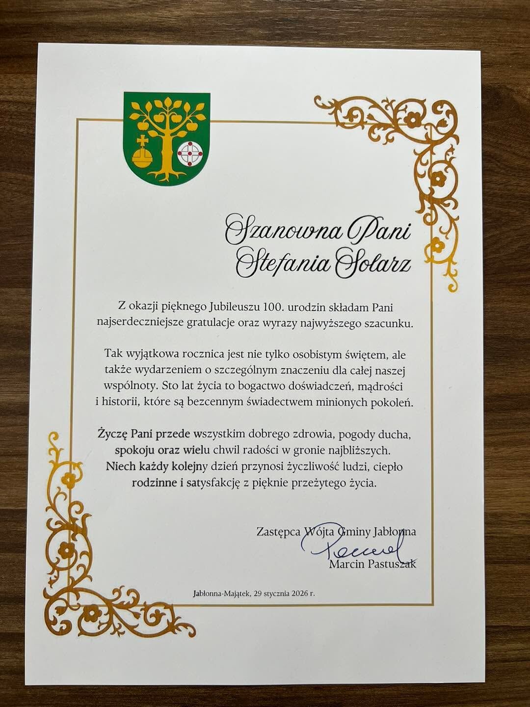 Dyplom okolicznościowy na papierze formatu A4 z dekoracyjną złotą bordiurą po prawej i lewej stronie oraz herbem w lewym górnym rogu.  Treść dyplomu (pisownia i układ zachowane): Szanowna Pani Stefania Solarz  Z okazji pięknego Jubileuszu 100. urodzin składam Pani najserdeczniejsze gratulacje oraz wyrazy najwyższego szacunku.  Tak wyjątkowa rocznica jest nie tylko osobistym świętem, ale także wydarzeniem o szczególnym znaczeniu dla całej naszej wspólnoty. Sto lat życia to bogactwo doświadczeń, mądrości i historii, które są bezcennym świadectwem minionych pokoleń.  Życzę Pani przede wszystkim dobrego zdrowia, pogody ducha, spokoju oraz wielu chwil radości w gronie najbliższych. Niech każdy kolejny dzień przynosi życzliwość ludzi, ciepło rodzinne i satysfakcję z pięknie przeżytego życia.  Zastępca Wójta Gminy Jabłonna (podpis) Marcin Pastuszak  Jabłonna-Majątek, 29 stycznia 2026 r.  Widoczne dekoracje: ornamenty roślinne w kolorze złotym w rogach i wzdłuż lewej krawędzi. Herb na zielonym tle przedstawia drzewo z owocami, po lewej symbol złotego oręża kościelnego, po prawej tarczę z czerwonym krzyżem na białym polu. Podpis nieczytelny ręcznie, nazwisko osoby podpisanej odczytane drukowanymi literami jako Marcin Pastuszak.