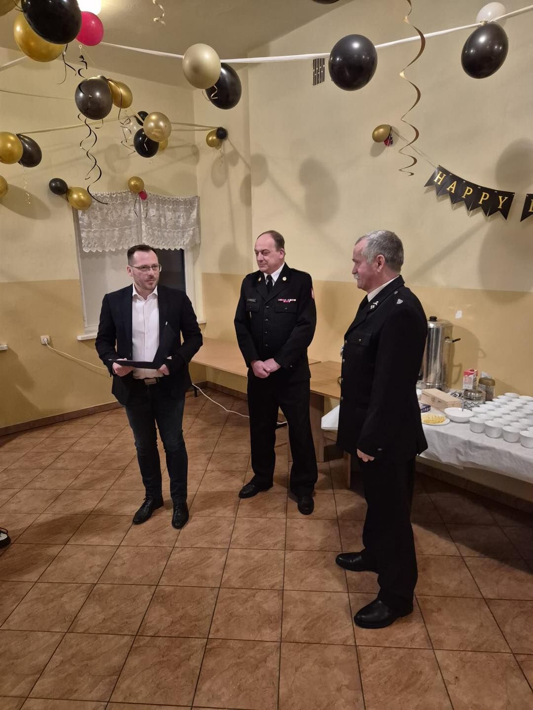 Na zdjęciu widać trzech mężczyzn stojących w sali. Po lewej stoi mężczyzna w ciemnej marynarce, białej koszuli i dżinsach, trzyma kartkę papieru jak podczas przemówienia. Obok niego dwóch mężczyzn w czarnych, galowych mundurach strażackich z odznaczeniami na piersi słucha go, zwróconych lekko w jego stronę. Za nimi po prawej stronie stoi stół przykryty białym obrusem z przygotowanym poczęstunkiem: rzędem białych kubków, dużym metalowym termosowym podgrzewaczem, dzbankami i talerzykami. Na ścianach wiszą czarno‑złote balony i girlanda z napisem „HAPPY”, tworząc uroczystą atmosferę.
