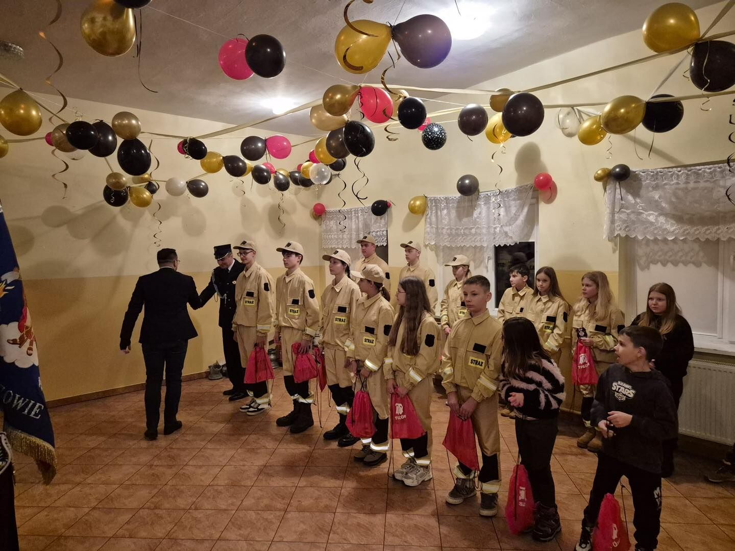 Grupa dzieci i nastolatków w jasnożółtych mundurach strażackich stoi w szeregu w ozdobionej sali. Mundury mają czarne naszywki na ramionach i żółte pasy odblaskowe na rękawach oraz nogawkach; na piersiach widoczny jest czarny napis „STRAŻ” na żółtym tle. Część młodzieży ma beżowe czapki z daszkiem, reszta jest bez nakrycia głowy. Większość osób w mundurach trzyma w dłoniach czerwone materiałowe worki–plecaki. Po lewej stronie dwóch dorosłych mężczyzn w ciemnych garniturach lub galowych mundurach rozmawia z pierwszym chłopcem w szeregu, jakby wręczali mu coś lub gratulowali. Po prawej stronie, pod ścianą, stoi kilka dziewcząt w takich samych mundurach, a przed nimi kilkoro dzieci w codziennych ubraniach, także z czerwonymi woreczkami. Na suficie i pod nim rozwieszona jest duża liczba balonów w kolorach złotym, czarnym, białym i czerwonym, z długimi zwisającymi wstążkami, co nadaje sali uroczysty, imprezowy charakter.