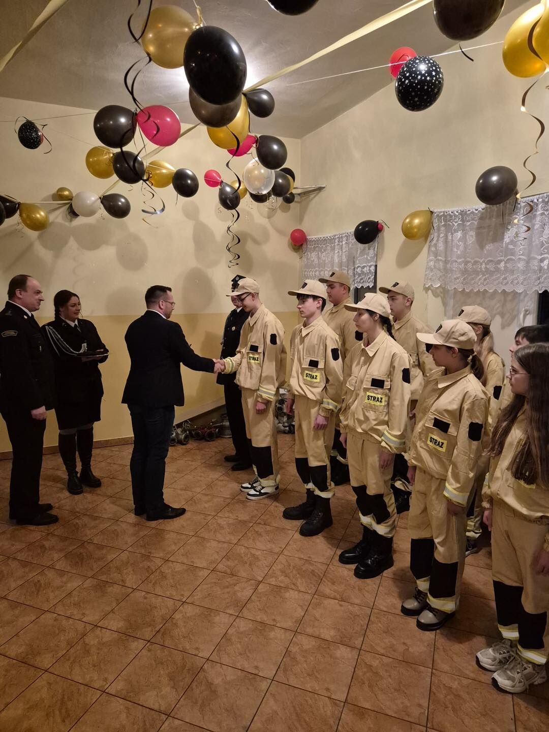 Na zdjęciu widać uroczystość w sali. Po prawej stronie stoi rząd młodzieży w jasnożółtych mundurach strażackich z naszywką „STRAŻ” na piersi i czarnymi wstawkami przy nogawkach; większość z nich ma beżowe czapki z daszkiem. Na środku mężczyzna w ciemnej marynarce ściska dłoń jednemu z chłopców w mundurze, jakby składał gratulacje lub wręczał wyróżnienie. Po lewej stronie stoją trzy osoby dorosłe w ciemnych, galowych strojach strażackich, obserwują sytuację. Nad głowami zwisają dziesiątki balonów w kolorach czarnym, złotym, czerwonym i białym, na długich, poskręcanych wstążkach, co tworzy odświętną atmosferę.