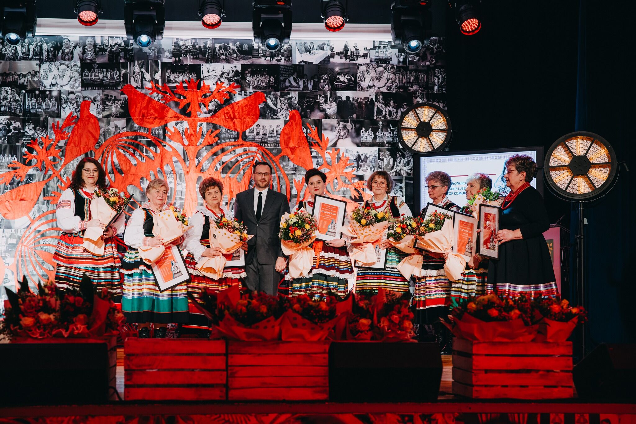 Grupa osób stojąca na scenie w trakcie uroczystości. Na środku stoi mężczyzna w ciemnym garniturze z białą koszulą i krawatem, po obu jego stronach jest kilka kobiet w kolorowych, ludowych strojach – mają białe koszule, ciemne gorsety i długie spódnice w poziome, wielobarwne pasy. Każda z kobiet trzyma duży bukiet kwiatów owinięty jasnym papierem oraz prostokątną ramę z dokumentem lub dyplomem w środku. Po prawej stronie stoi jeszcze jedna kobieta w czarnej sukience z kolorowym, pasowym wykończeniem u dołu oraz czerwonym naszyjnikiem; trzyma w rękach ramę i kopertę. Przed grupą, na przodzie sceny, ustawione są drewniane skrzynki wypełnione czerwonymi kwiatami. W tle widać duży, dekoracyjny motyw wycinanki w intensywnym czerwonym kolorze z ptakami i roślinnymi kształtami oraz czarno‑białą ścianę z wieloma małymi zdjęciami. Po bokach sceny stoją duże, okrągłe lampy świecące ciepłym światłem.