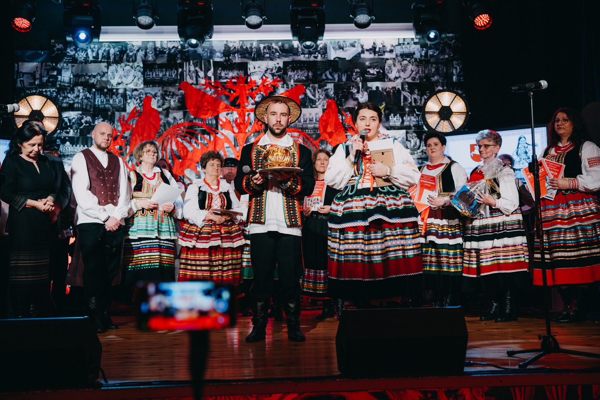 Na scenie stoi grupa osób biorących udział w uroczystości. W centrum jest mężczyzna w ludowym stroju: ma kapelusz ze słomy, białą koszulę, czarną, bogato haftowaną kamizelę i spodnie z wysokimi butami; trzyma mikrofon oraz okrągłą, złotą statuetkę lub puchar na tacy. Obok niego, po prawej, stoi kobieta w kolorowym stroju ludowym z rozkloszowaną spódnicą w poziome pasy; mówi do mikrofonu i trzyma teczkę lub nagrodę pod pachą. Za nimi i obok nich stoją inne kobiety w podobnych, pasiastych strojach ludowych, trzymają dyplomy, programy i upominki w opakowaniach. Po lewej stronie widoczny jest mężczyzna w białej koszuli i bordowym, ludowym kaftanie oraz kobieta w czarnej sukience. Tło sceny jest takie samo jak wcześniej: czerwony motyw wycinanki z ptakami na ścianie pełnej czarno‑białych zdjęć oraz duże, okrągłe lampy. Na dole, z przodu, rozmazany prostokątny kształt – prawdopodobnie telefon nagrywający wydarzenie.
