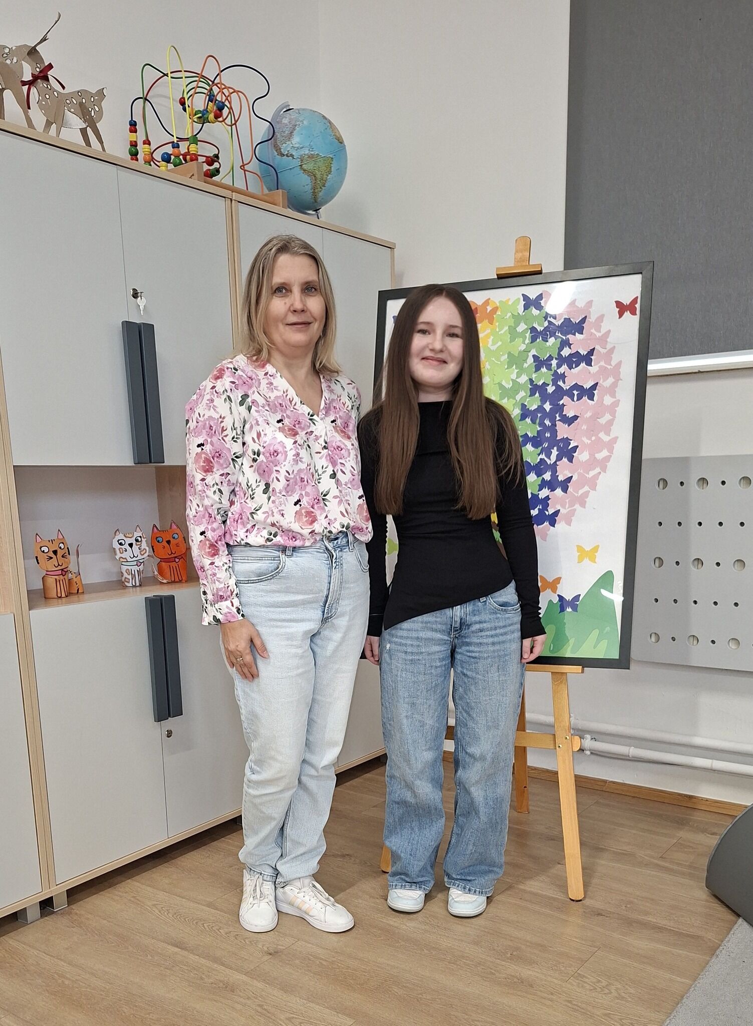 Dwie osoby stojące obok siebie w pomieszczeniu, przy sztaludze z kolorowym obrazem motyli.  Po lewej stronie stoi dorosła kobieta o jasnej cerze, blond włosach do ramion i neutralnym wyrazie twarzy. Ma na sobie jasną koszulę w różowy kwiatowy wzór, jasnoniebieskie dżinsy i białe buty sportowe.  Po prawej stoi nastolatka o jasnej cerze i długich, prostych, brązowych włosach. Ma na sobie czarną, dopasowaną bluzkę z długim rękawem, niebieskie dżinsy i jasne buty.  Za nimi, po prawej, stoi sztaluga z oprawioną pracą przedstawiającą wiele kolorowych motyli (m.in. różowe, fioletowe, niebieskie, zielone, żółte) ułożonych w gęsty wzór.  W tle po lewej widać jasne szafki; na górze stoją drewniane figurki jeleni, kolorowa zabawka z drucikami i koralikami oraz globus. Na półce są małe figurki kotów. Po prawej stronie jest fragment ściany z jasną tablicą z okrągłymi otworami i grzejnikiem.