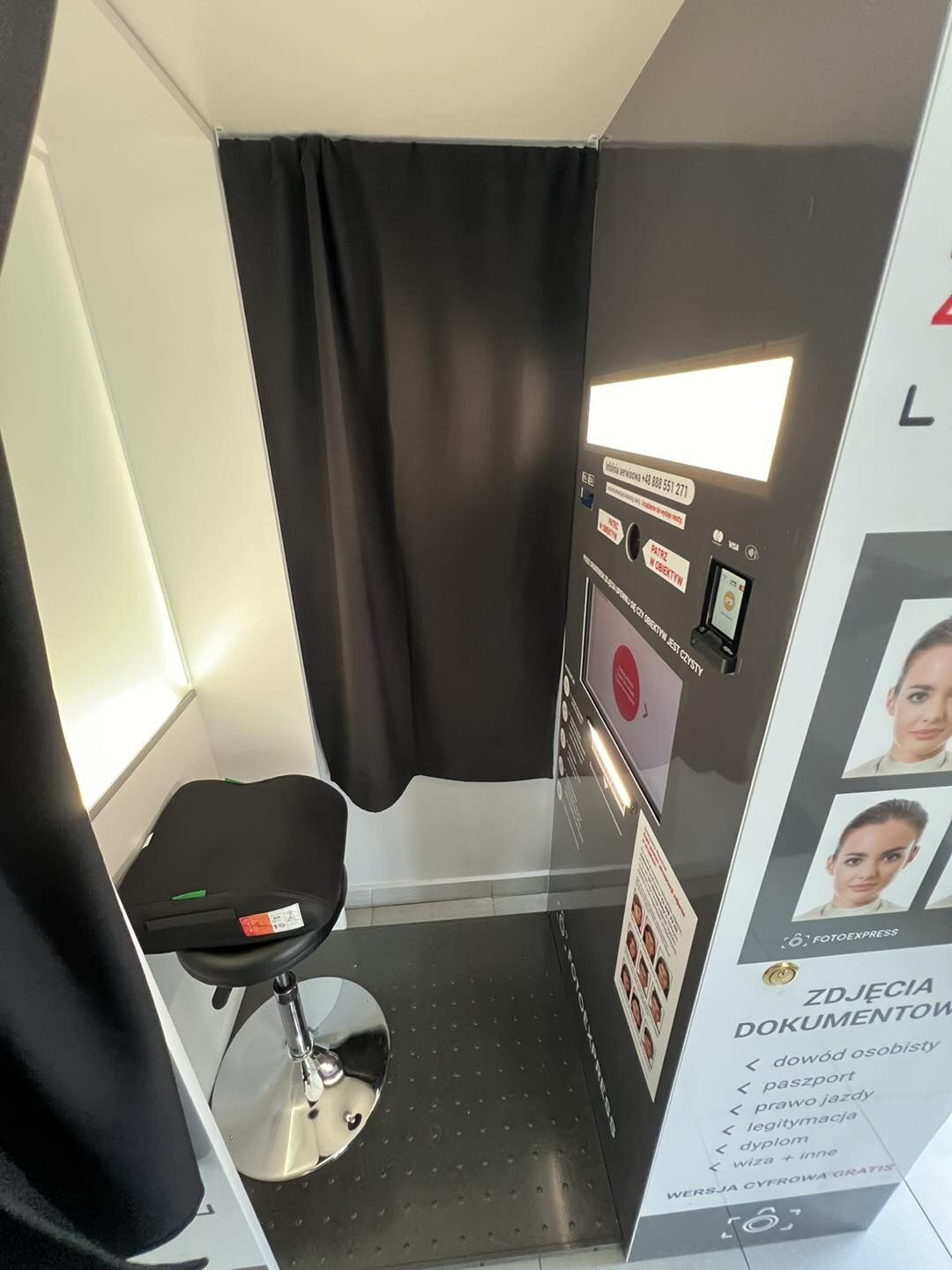 Wnętrze fotobudki do zdjęć.  Po lewej stronie jest jasny panel świetlny (prostokątny), a obok czarne siedzisko na metalowej, regulowanej nodze z okrągłą chromowaną podstawą.  Na wprost, przy tylnej ścianie, wisi czarna zasłona.  Po prawej jest pionowy panel sterujący urządzenia: u góry świecący prostokątny ekran/podświetlany obszar, poniżej gniazda/otwory (w tym okrągły otwór) i naklejki z instrukcjami, dalej większy ekran dotykowy, a niżej wydrukowana instrukcja obrazkowa. Po prawej stronie panelu jest też mały czytnik/terminal płatniczy.  Podłoga jest czarna, z regularnymi małymi wypustkami (antypoślizgowa).