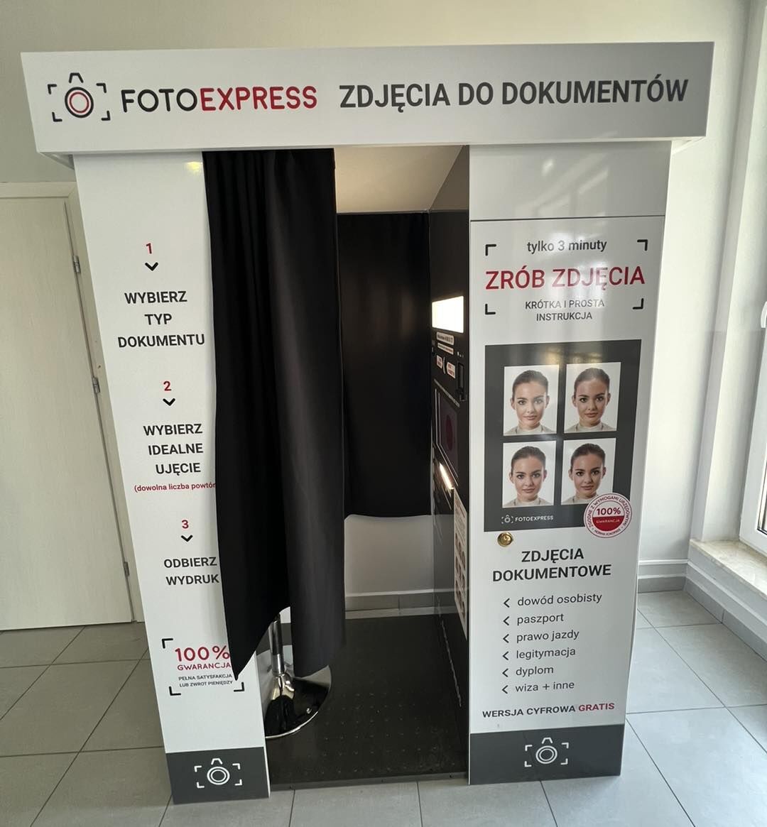 Biała fotobudka do zdjęć dokumentowych z czarną zasłoną wejściową pośrodku.  Na górnym szyldzie jest napis: „FOTOEXPRESS ZDJĘCIA DO DOKUMENTÓW”.  Lewa strona budki ma instrukcję w trzech krokach: „1 WYBIERZ TYP DOKUMENTU” „2 WYBIERZ IDEALNE UJĘCIE (dowolna liczba powtór…)” (końcówka ucięta na zdjęciu) „3 ODBIERZ WYDRUK” Niżej jest znak „100% GWARANCJA” i mniejszy tekst, który jest zbyt mały, by odczytać w całości.  Prawa strona budki ma napis: „tylko 3 minuty ZRÓB ZDJĘCIA” oraz „KRÓTKA I PROSTA INSTRUKCJA”. Poniżej jest panel z czterema przykładowymi zdjęciami twarzy kobiety oraz okrągła pieczęć z „100%” (reszta drobnym drukiem). Niżej: „ZDJĘCIA DOKUMENTOWE” i lista: „dowód osobisty, paszport, prawo jazdy, legitymacja, dyplom, wiza + inne”. Na dole: „WERSJA CYFROWA GRATIS”.  W środku, za zasłoną, widać czarną podłogę z kropkami i metalowy słupek. Po prawej wewnątrz jest panel urządzenia z ekranem i elementami sterującymi.