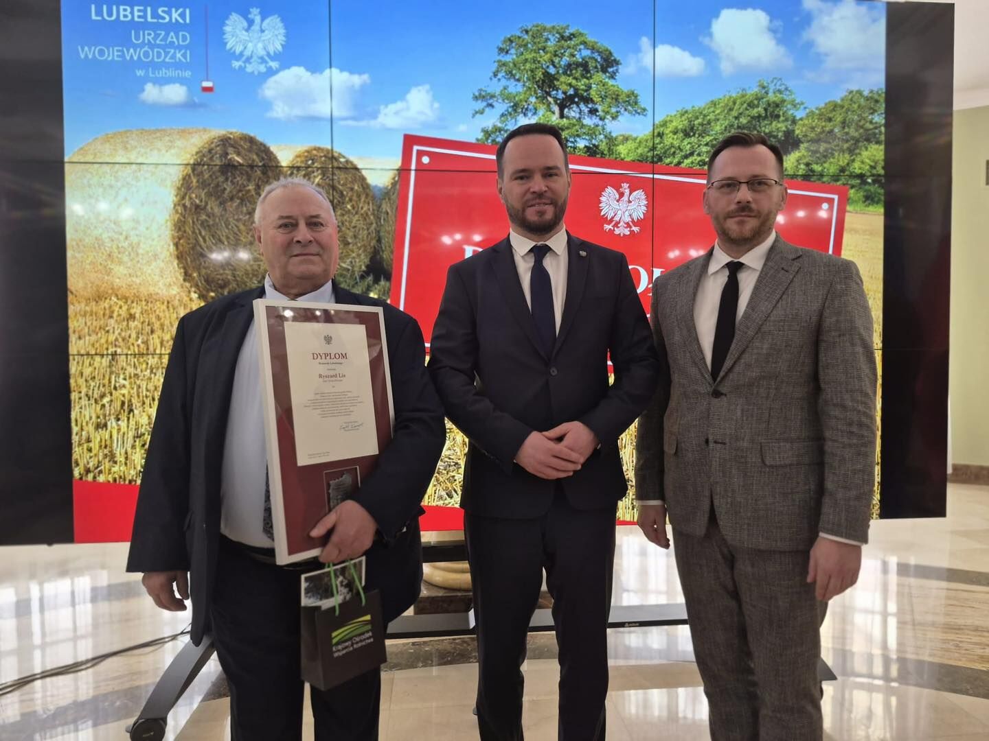 Trzech mężczyzn w garniturach stojących przed dużym ekranem z grafiką pola ze zbożem i belami siana oraz polskimi symbolami państwowymi.  W tle, w lewym górnym rogu, jest napis: „LUBELSKI URZĄD WOJEWÓDZKI w Lublinie” oraz biały orzeł. Mężczyzna po lewej (starszy, jasna skóra, siwe włosy) trzyma w ramie dokument z dużym tytułem „DYPLOM”; na dokumencie widoczne jest imię i nazwisko zaczynające się od „Ryszard Li…”. Trzyma też ciemną torbę prezentową z zielonymi elementami i tekstem, którego nie da się w całości odczytać. Mężczyzna pośrodku (jasna skóra, ciemne włosy, krótki zarost) ma granatowy garnitur i krawat; stoi z dłońmi złożonymi z przodu. Mężczyzna po prawej (jasna skóra, krótkie ciemne włosy, zarost, okulary) ma szary garnitur i czarny krawat.  Na ekranie za nimi widać także dużą czerwoną planszę z białym orłem; część tekstu na niej jest zasłonięta i nieczytelna.