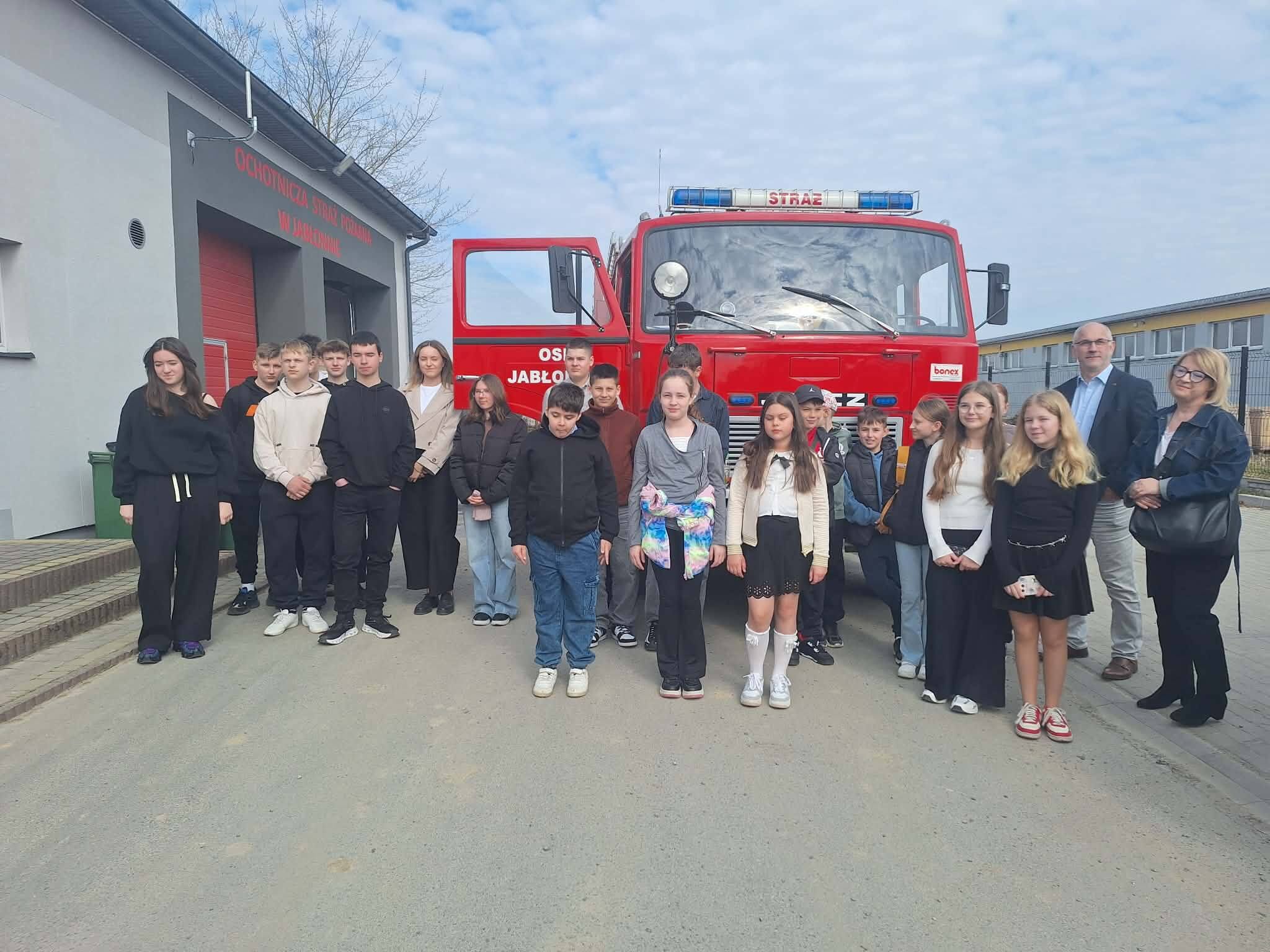 Grupa dzieci i dorosłych stojąca przed czerwonym wozem strażackim przy budynku remizy. Na zdjęciu widać około 20 osób ustawionych w kilku rzędach; większość to dzieci i nastolatki, a po prawej stronie stoją też dwie osoby dorosłe (mężczyzna w marynarce i koszuli oraz kobieta w okularach i ciemnym stroju). W centrum kadru znajduje się duży czerwony samochód strażacki z niebieskimi lampami na dachu; na górze jest napis „STRAŻ”, a na przodzie widoczne jest duże białe „JELCZ”. Jedne drzwi kabiny są otwarte.  Po lewej stronie stoi budynek z czerwonymi bramami garażowymi; nad nimi znajduje się czerwony napis „OCHOTNICZA STRAŻ POŻARNA”, a poniżej drugi wiersz z nazwą miejscowości zaczynającą się od „JABŁO…”. Osoby są ubrane głównie w codzienne ubrania w ciemnych kolorach (bluzy, spodnie, kurtki), część dziewczynek ma jasne bluzki i spódnice; jedna dziewczynka z przodu ma kolorową kurtkę przewieszoną na rękach. Tło to jasne, lekko zachmurzone niebo oraz fragmenty zabudowań po prawej.