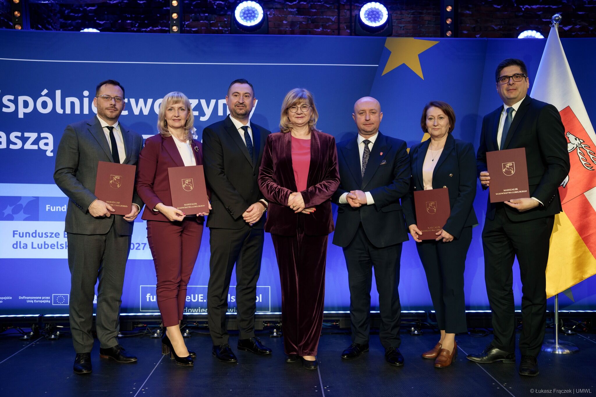 Grupa siedmiu osób stojących na scenie na tle niebieskiej ścianki z motywami Unii Europejskiej. To zdjęcie z wydarzenia oficjalnego.  Od lewej do prawej stoją cztery osoby trzymające bordowe teczki/dyplomy z herbem: mężczyzna w szarym garniturze i okularach, kobieta w bordowym garniturze, kobieta w granatowym żakiecie oraz mężczyzna w ciemnym garniturze i okularach (ten po prawej). Trzy osoby w środku nie trzymają teczek: mężczyzna w ciemnym garniturze, kobieta w bordowym welurowym komplecie z różową bluzką, oraz łysy mężczyzna w ciemnym garniturze.  Na tle widać dużą żółtą gwiazdę (symbol UE) i fragmenty napisów po polsku, m.in. „Fundusze … dla Lubels…” oraz „Dofinansowane przez Unię Europejską”. Po prawej stronie stoi flaga w barwach białej i czerwonej z herbem. Oświetlenie sceniczne jest mocne, z widocznymi lampami nad osobami.