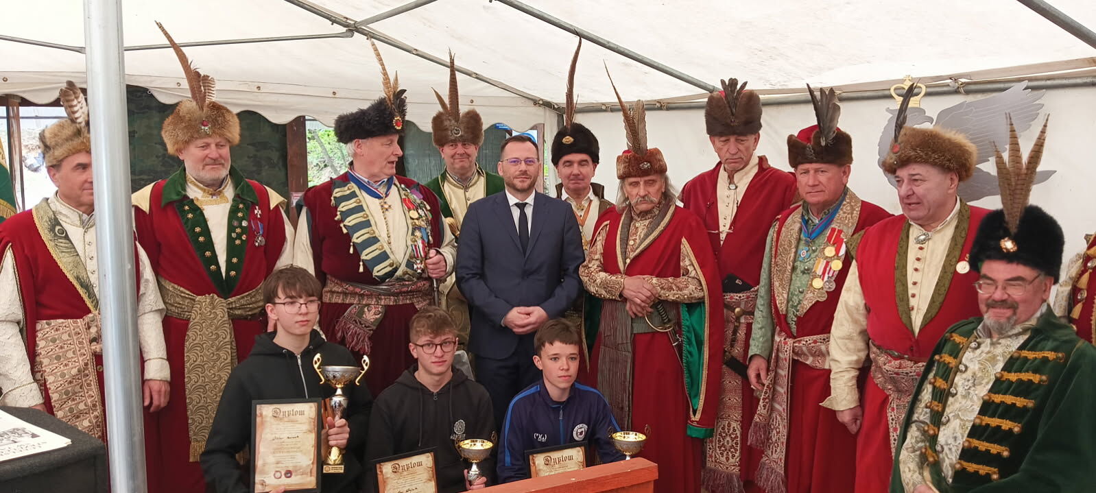 Grupa mężczyzn w strojach historycznych oraz kilku chłopców z pucharami i dyplomami. Na zdjęciu widać kilkunastu dorosłych mężczyzn stojących w dwóch rzędach pod białym namiotem. Większość z nich ma na sobie bogato zdobione szlacheckie kontusze, głównie czerwone i zielone, przepasane pasami z dekoracyjnymi wzorami. Na głowach mają futrzane czapki z długimi piórami sterczącymi pionowo do góry. Część mężczyzn ma na piersiach medale i ordery, niektórzy również ozdobne łańcuchy. Pośrodku tylnego rzędu stoi mężczyzna w nowoczesnym, ciemnogranatowym garniturze, białej koszuli i krawacie, wyróżniający się na tle historycznych strojów. Przed dorosłymi, w przednim rzędzie, siedzi trzech nastolatków i jeden młodszy chłopiec. Chłopcy ubrani są współcześnie, głównie w bluzy sportowe lub dresowe. Trzech z nich trzyma złote puchary, a co najmniej dwóch trzyma dyplomy w ozdobnych ramkach lub teczkach z napisem „Dyplom” u góry. Przed chłopcami stoi niski drewniany podest lub ławka w kolorze pomarańczowo-brązowym. Mimika większości osób jest poważna lub lekko uśmiechnięta, wszyscy ustawieni są tak, jak do zdjęcia pamiątkowego po jakimś konkursie lub uroczystości.