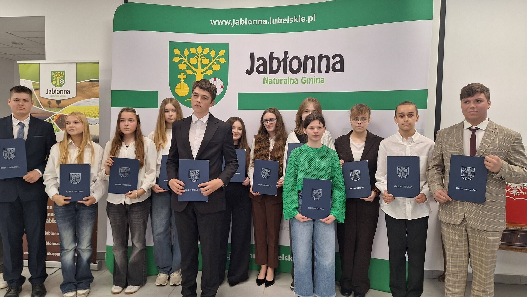 Grupa kilkunastu nastolatków i nastolatek stoi w rzędzie, trzymając granatowe teczki z herbem i napisem „Gmina Jabłonna”. Wszyscy są odświętnie ubrani – większość ma białe koszule, część garnitury lub eleganckie marynarki, jedna dziewczyna wyróżnia się zielonym, grubym swetrem i szerokimi dżinsami. Dzieci i młodzież stoją na tle dużej ścianki z herbem i napisem „Jabłonna Naturalna Gmina” oraz adresem internetowym „www.jablonna.lubelskie.pl” na zielonym pasku u góry. Po lewej stronie widoczny jest dodatkowy stojący baner promocyjny z logotypem Jabłonny, po prawej fragment godła Polski na czerwonym tle. Osoby patrzą przed siebie z poważnymi lub lekkimi uśmiechami, ułożone równo w jednym rzędzie, co nadaje scenie oficjalny, uroczysty charakter – wygląda to jak rozdanie nagród lub stypendiów.