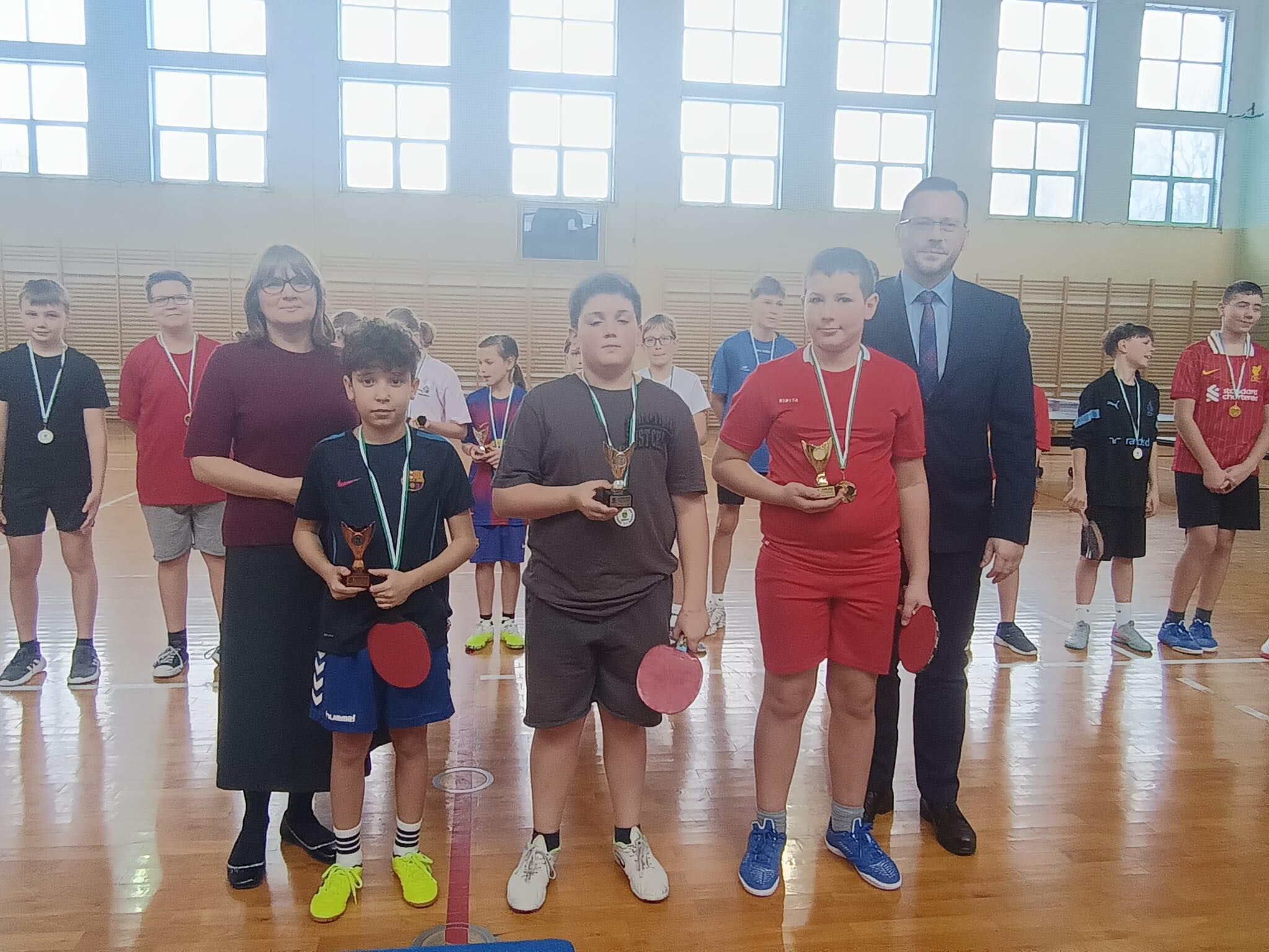 Zdjęcie z sali gimnastycznej podczas wręczania nagród w tenisie stołowym.  Na pierwszym planie stoi trzech chłopców z medalami: Po lewej chłopiec w ciemnej koszulce (z herbem Barcelony) i niebieskich spodenkach, trzyma mały puchar i ma w drugiej ręce czerwoną rakietkę; ma jaskrawożółte buty. W środku chłopiec w ciemnoszarym stroju, trzyma puchar i czerwoną rakietkę. Po prawej chłopiec w czerwonym stroju sportowym, trzyma puchar i w drugiej ręce czerwoną rakietkę; ma niebieskie buty.  Za nimi stoją dwoje dorosłych: Po lewej kobieta w okularach, w bordowym swetrze i długiej ciemnej spódnicy. Po prawej mężczyzna w garniturze, w okularach.  W tle widać innych uczestników z medalami oraz wysokie okna i drabinki na ścianie.