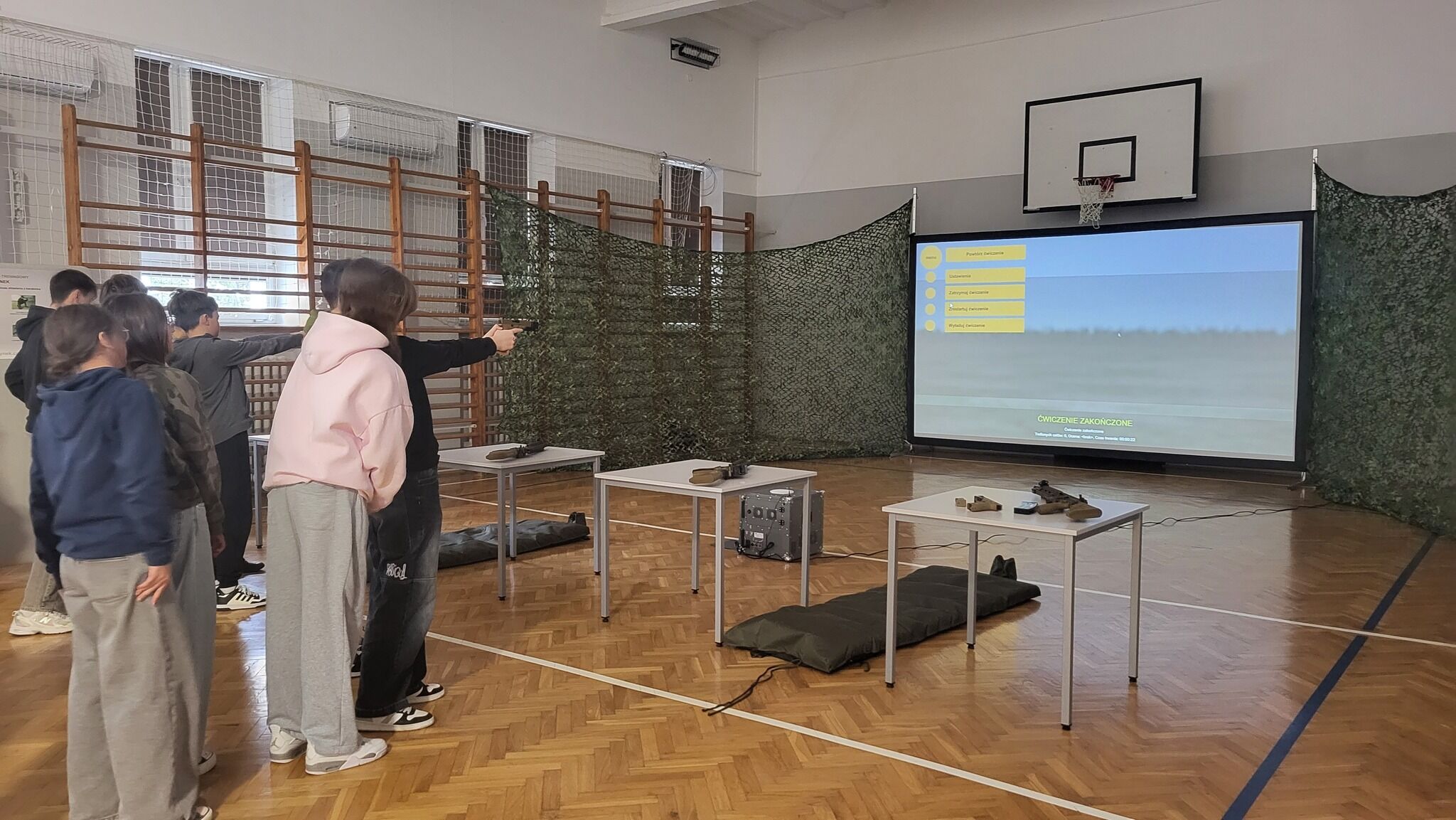 Grupa os&oacute;b w sali gimnastycznej korzysta z symulatora strzeleckiego. Przed nimi ekran z obrazem krajobrazu. Na stolikach leżą symulacyjne karabiny. W tle wysoka siatka i kosz do koszyk&oacute;wki.