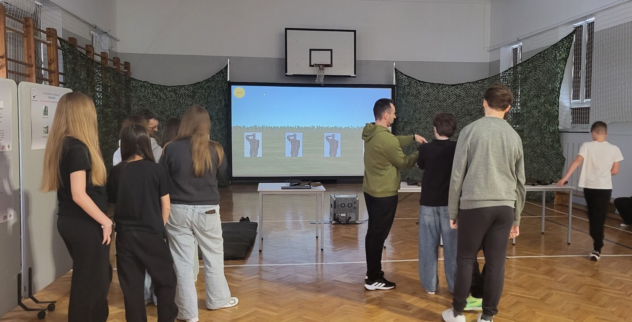 Grupa os&oacute;b stoi w sali gimnastycznej przed ekranem, na kt&oacute;rym wyświetlany jest symulator strzelnicy z tarczami. Na ścianach zawieszone są siatki maskujące. Osoby są w trakcie ćwiczeń lub instruktażu.