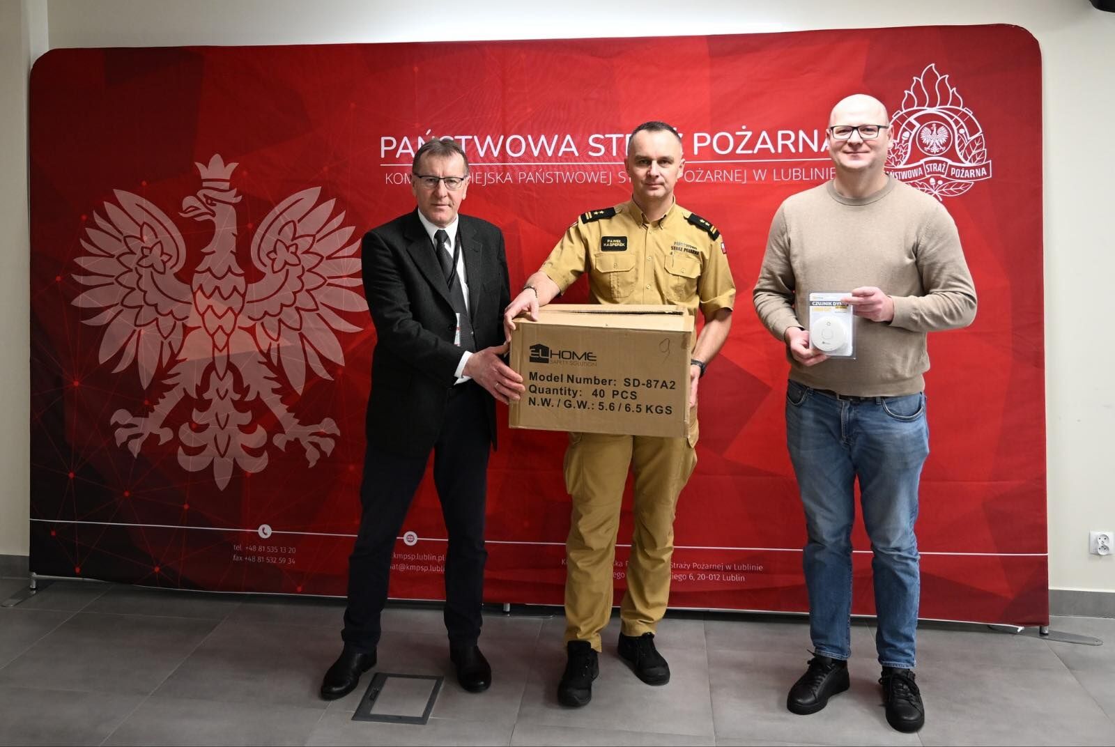 Trzech mężczyzn stojących przed czerwonym tłem z napisem „PAŃSTWOWA STRAŻ POŻARNA” oraz godłami (biały orzeł po lewej i emblemat straży po prawej). Po lewej stoi mężczyzna w czarnym garniturze, białej koszuli i czarnym krawacie, w okularach. W środku stoi mężczyzna w piaskowym mundurze strażackim, trzyma duże kartonowe pudło z nadrukiem „ELHOME”, „Model Number: SD-87A2”, „Quantity: 40 PCS”, „N.W./G.W.: 5.6/6.5 KGS”. Po prawej stoi mężczyzna w jasnym swetrze i niebieskich dżinsach, w okularach, trzyma zapakowane urządzenie z napisem „CZUJNIK DYMU” na opakowaniu.