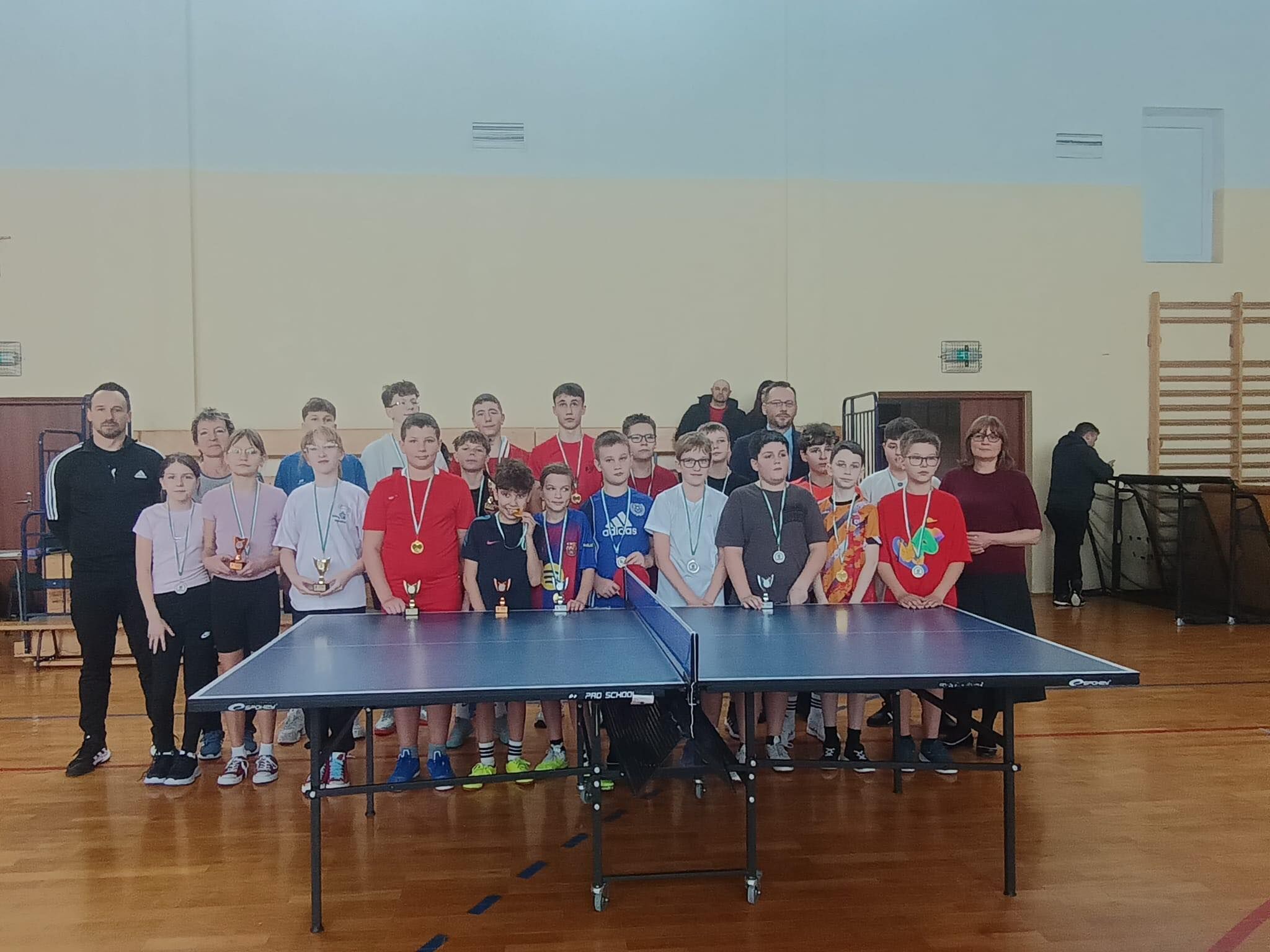 Grupa dzieci i dorosłych stojąca w sali gimnastycznej za stołem do tenisa stołowego. Widać około dwudziestu kilku osób, głównie dzieci i nastolatków, ustawionych w dwóch rzędach. Wiele osób ma na szyi medale na wstążkach, a kilka trzyma małe puchary. Na pierwszym planie stoi niebieski stół do ping-ponga z siatką pośrodku, na metalowych nogach z kółkami. Po lewej stronie stoi dorosły mężczyzna w czarnym dresie, a po prawej dorosła kobieta w ciemnym swetrze; w tle jest jeszcze kilku dorosłych. W tle są jasne ściany sali, drabinki gimnastyczne po prawej oraz sprzęt sportowy złożony przy ścianie.