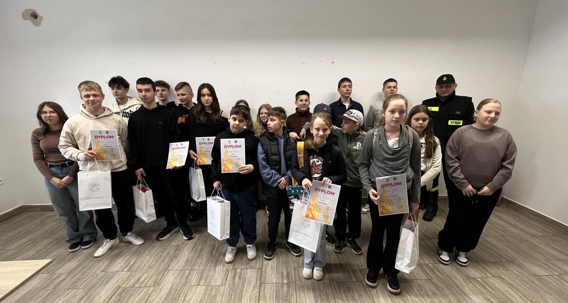 Grupa nastolatków i kilkorga młodszych dzieci ustawiona w dwóch rzędach przy białej ścianie w pustym pomieszczeniu z szarobrązową, drewnopodobną podłogą. Wiele osób trzyma białe torby prezentowe oraz dyplomy.  Po lewej stronie stoi dziewczyna w okularach, w brązowym swetrze i ciemnych spodniach. Obok niej chłopak w jasnej bluzie z kapturem trzyma dyplom; na dyplomie wyraźnie widać duży napis „DYPLOM”. Dalej w szeregu stoją głównie chłopcy w ciemnych bluzach i kurtkach, a kilka osób w tle jest częściowo zasłoniętych.  W środkowej części widać kilkoro młodszych dzieci z dyplomami „DYPLOM” i białymi torbami. Jedna dziewczynka z przodu, w ciemnej bluzie, też trzyma dyplom. Obok niej stoi chłopiec w zielonej bluzie z nadrukiem i czapce z daszkiem.  Po prawej stronie z przodu stoi dziewczyna w szarym szalu/kominie i ciemnych spodniach, trzyma dyplom „DYPLOM” oraz białą torbę. Najbardziej z prawej stoi dziewczyna w szaro-brązowej bluzie i ciemnych spodniach, z rękami splecionymi z przodu.  W tle po prawej stronie stoi dorosły mężczyzna w czarnym mundurze z naszywką „STRAŻ” i czarną czapką. Na ścianie wysoko po lewej widać małą, nieregularną plamę lub ubytek.