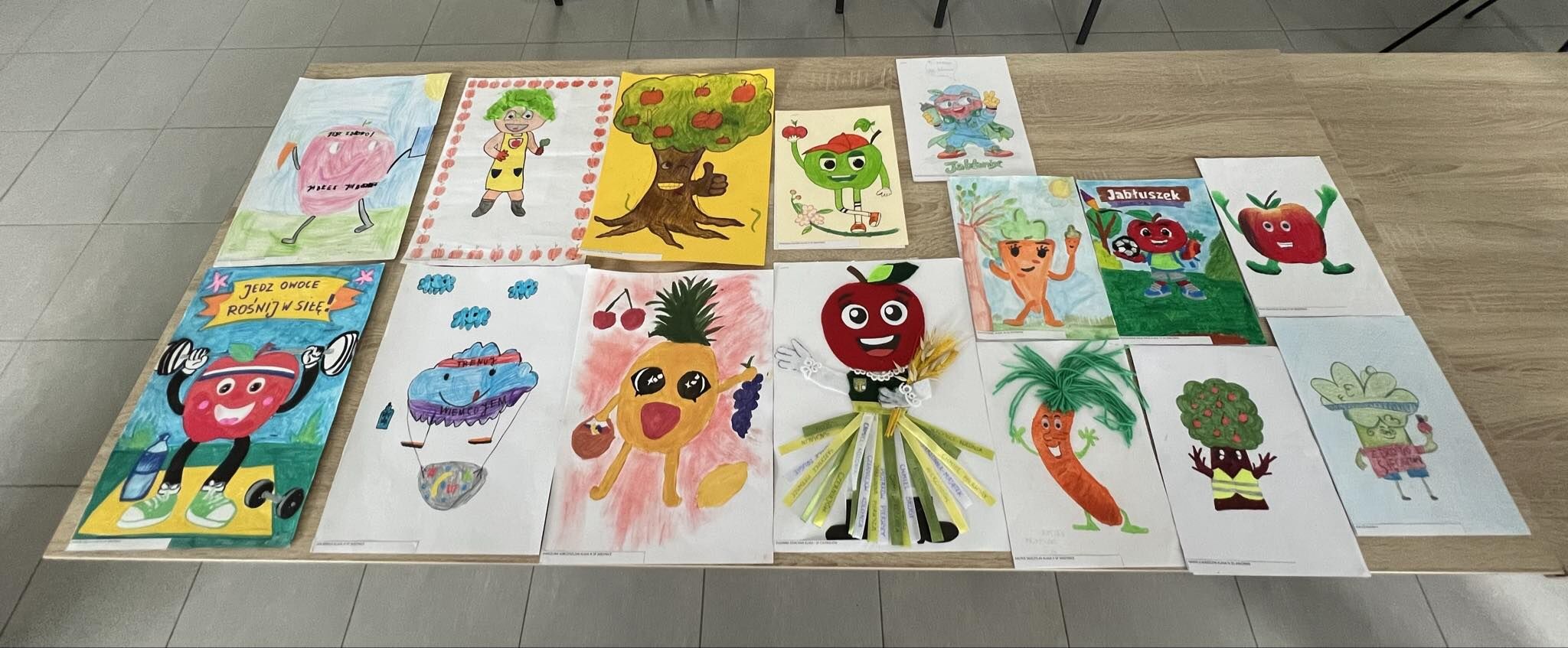 Zestaw dziecięcych rysunków i prac plastycznych rozłożonych na dużym drewnianym stole w dwóch rzędach. To głównie uśmiechnięte owoce i warzywa z oczami, rękami i nogami; jedna praca ma elementy przestrzenne z papierowych pasków.  W górnym rzędzie, od lewej: Rysunek różowego owocu z rękami i nogami na jasnoniebieskim tle; na owocu jest napis, ale jest nieczytelny. Postać dziecka z zielonymi włosami w żółtym stroju, w ramce z czerwonych kropek. Drzewo z twarzą i uniesionym kciukiem, na koronie czerwone owoce. Zielone jabłko/owoc w czapce, z uniesioną ręką; obok małe kwiatki. Kartka z postacią w niebieskim stroju; podpis pod rysunkiem jest nieczytelny. Marchewka z twarzą, z uniesionymi rękami; obok słoneczko. Rysunek z tytułem „Jabłuszek”; czerwone jabłko jako piłkarz z piłką. Czerwone jabłko z rękami i nogami na białym tle.  W dolnym rzędzie, od lewej: Plakat z napisem „JEDZ OWOCE ROSNIJ W SILE!” z uśmiechniętym czerwonym owocem jako ciężarowcem. Niebiesko-fioletowy owoc/postać z napisem „TRENING” na opasce; u dołu kosz z kolorowymi elementami. Żółty ananas z twarzą; obok wiśnie i kiść winogron. Czerwone jabłko z dużymi oczami i uśmiechem; pod nim doczepione papierowe paski jak spódnica, z drobnymi napisami (nieczytelne). Marchewka z zielonym „pióropuszem” liści, z uśmiechem. Brokuł/drzewko z twarzą i rękami. Zielony owoc w kapeluszu, trzyma coś małego; napisy są nieczytelne.
