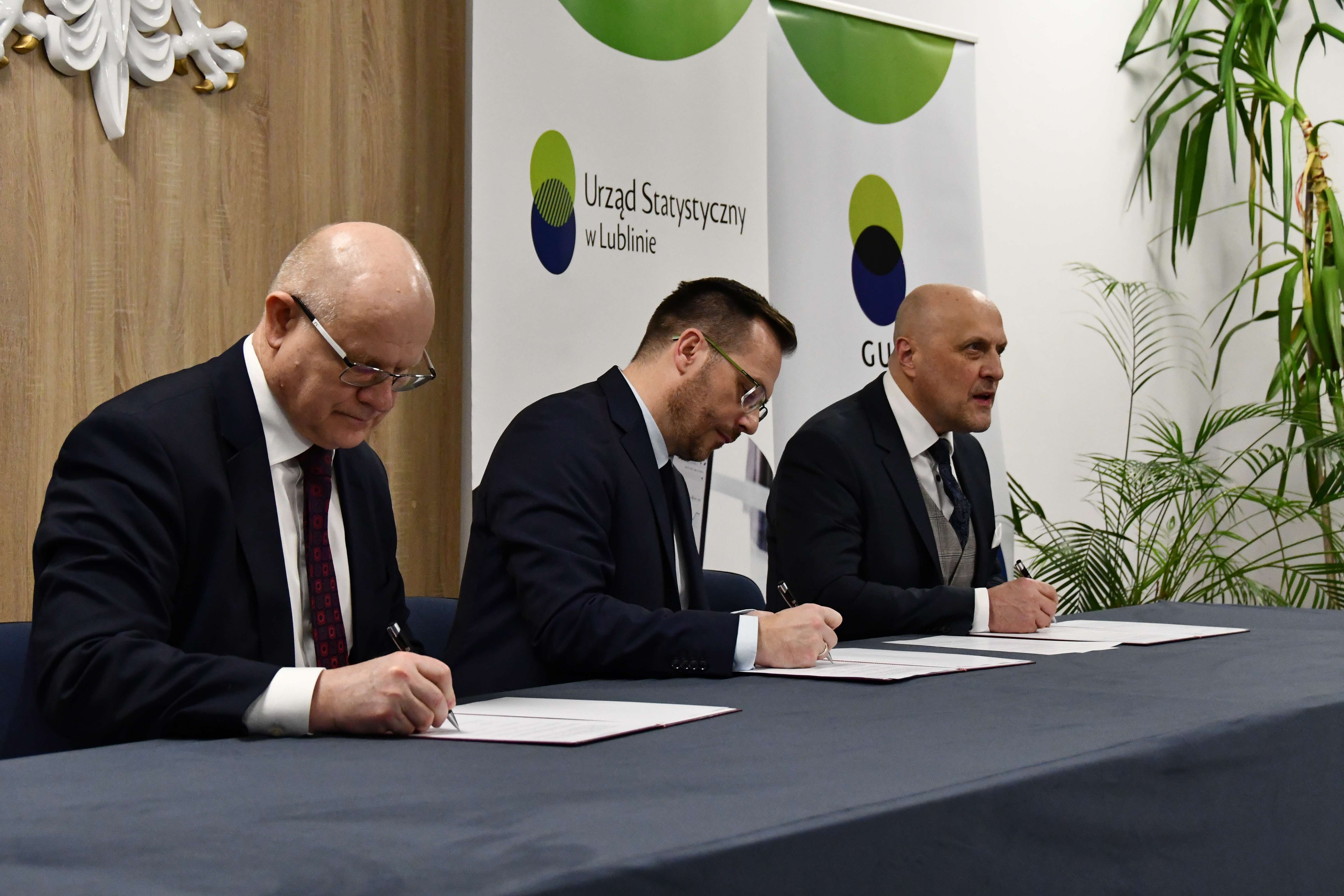 Trzech mężczyzn w garniturach siedzi przy długim stole i podpisuje dokumenty leżące przed nimi w bordowych teczkach. Po lewej siedzi łysy mężczyzna o jasnej karnacji w granatowym garniturze, białej koszuli i bordowym krawacie we wzór, w okularach. Pośrodku siedzi mężczyzna o jasnej karnacji z krótkimi brązowymi włosami i brodą, w granatowym garniturze, białej koszuli, ciemnym krawacie i okularach. Po prawej siedzi łysy mężczyzna o jasnej karnacji w granatowym garniturze, jasnoszarej kamizelce, białej koszuli i granatowym krawacie z wzorem oraz poszetką. W tle stoją pionowe banery z logo i napisem „Urząd Statystyczny w Lublinie” oraz „GUS”, a po prawej stronie widoczne są wysokie zielone rośliny.