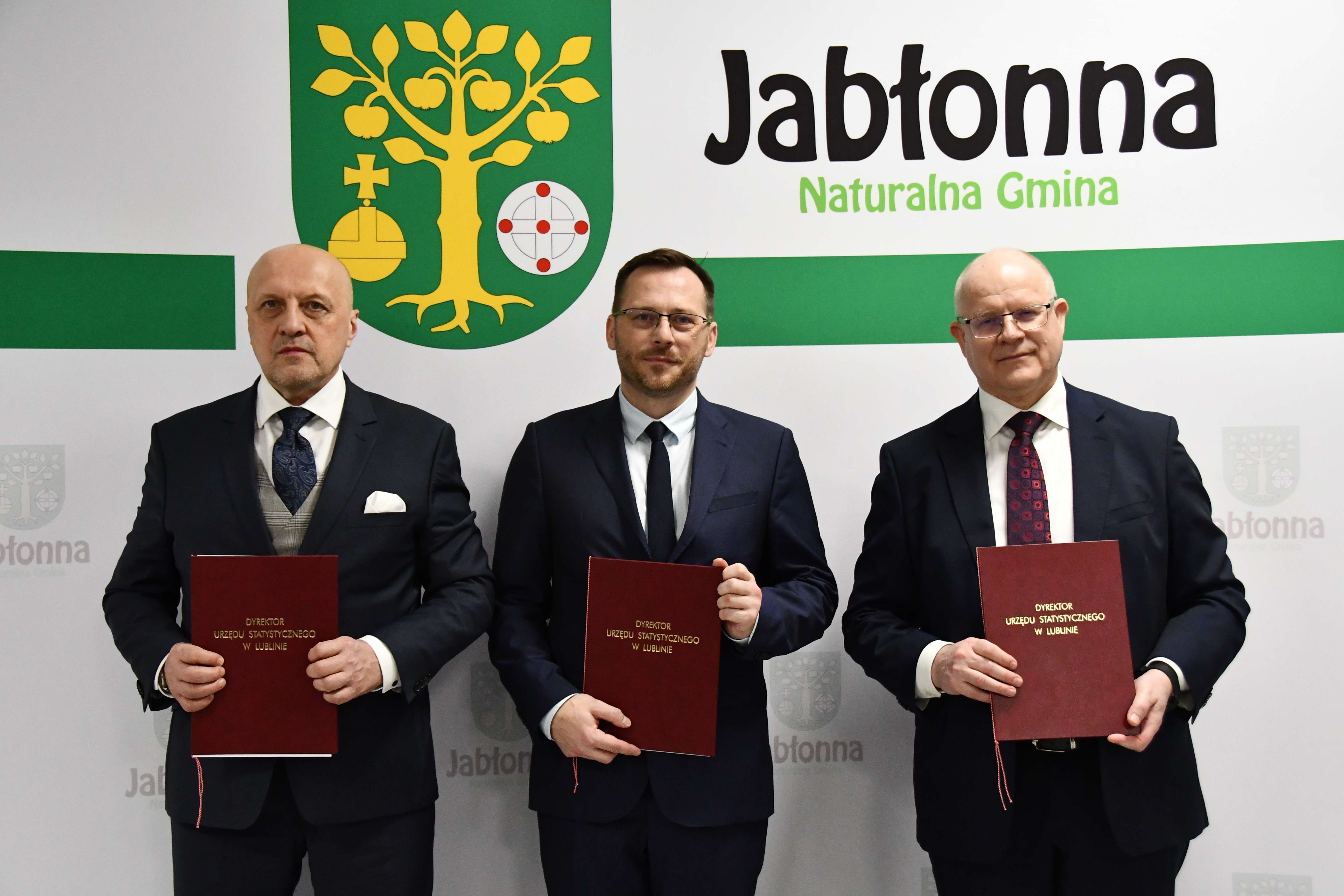 Trzech mężczyzn w garniturach trzymających bordowe teczki. Po lewej stoi łysy mężczyzna o jasnej karnacji, w ciemnym granatowym garniturze, jasnoszarej kamizelce, białej koszuli i granatowym krawacie z wzorem; z kieszonki wystaje biały poszetka. Pośrodku stoi mężczyzna o jasnej karnacji, z krótkimi brązowymi włosami i brodą, w granatowym garniturze, białej koszuli i ciemnym krawacie; ma okulary w cienkich oprawkach. Po prawej jest mężczyzna o jasnej karnacji, łysy, w granatowym garniturze, białej koszuli i bordowym krawacie z wzorem; nosi okulary. Każdy z nich trzyma przed sobą bordową teczkę z złotym napisem „DYREKTOR URZĘDU STATYSTYCZNEGO W LUBLINIE”. W tle widoczne jest duże zielono-żółte godło z drzewem jabłoni z owocami oraz napis „Jabłonna Naturalna Gmina” na jasnym tle.