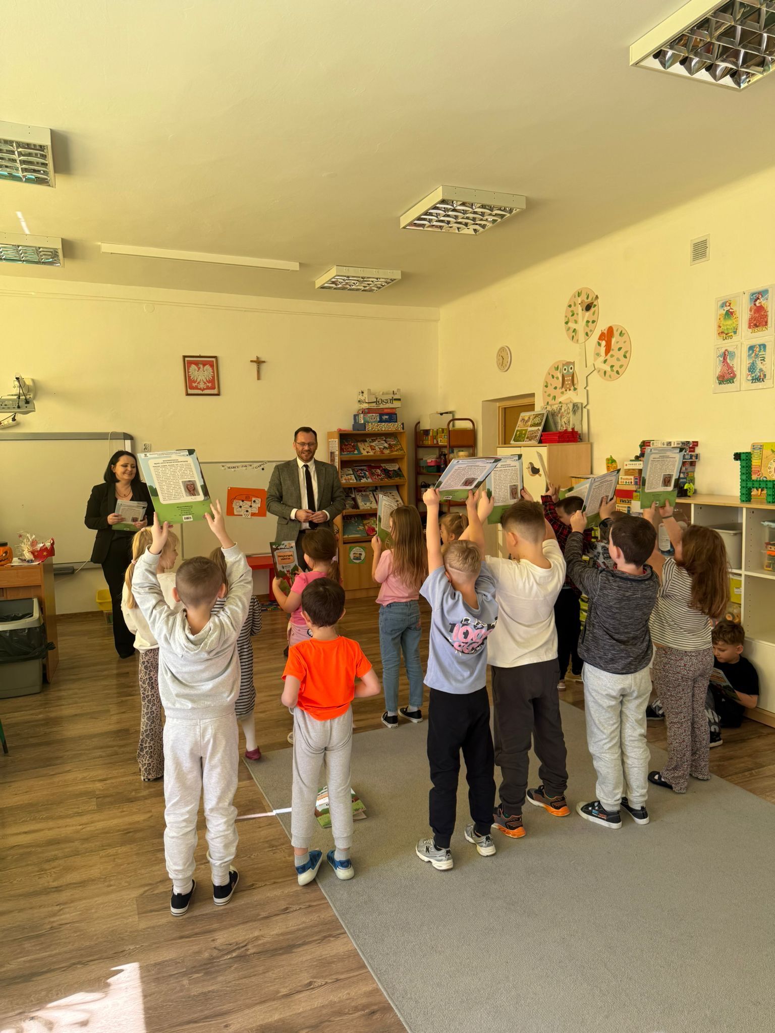 Sala przedszkolna. Grupa dzieci stoi na dywanie, zwrócona plecami do aparatu, i trzyma wysoko w górze otwarte książki lub zeszyty.  Z przodu, przy ścianie, stoi dorosły mężczyzna w garniturze i obok niego dorosła kobieta w ciemnym stroju; oboje wyglądają na prowadzących zajęcia. Na ścianie widać godło Polski i krzyż. W tle są regały z książkami i grami oraz niskie szafki z zabawkami.