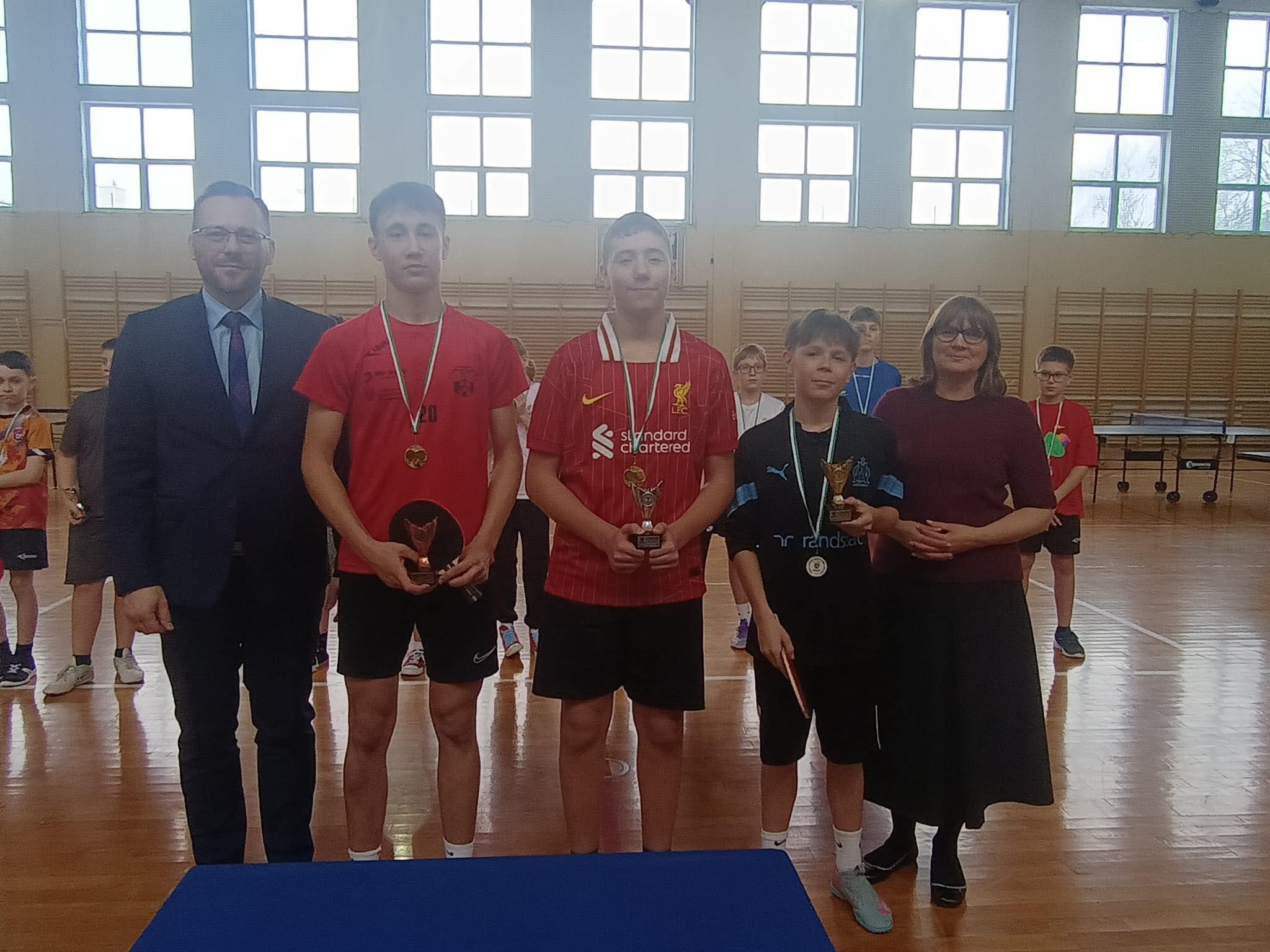 Pięć osób stoi w sali gimnastycznej, najpewniej podczas wręczania nagród.  Od lewej: Mężczyzna w garniturze (ciemny), biała koszula, krawat, okulary. Wysoki chłopak w czerwonej koszulce i czarnych spodenkach, ma medal na szyi i trzyma nagrodę. Chłopak w czerwonej koszulce w prążki (Liverpool) i czarnych spodenkach, ma medal i trzyma puchar. Niższy chłopak w czarnej koszulce i czarnych spodenkach, ma medal i trzyma puchar; w drugiej ręce trzyma rakietkę do tenisa stołowego. Kobieta w ciemnym swetrze i długiej ciemnej spódnicy, w okularach.  W tle widać innych uczestników (dzieci/nastolatków), drabinki na ścianie i stoły do ping-ponga po prawej.