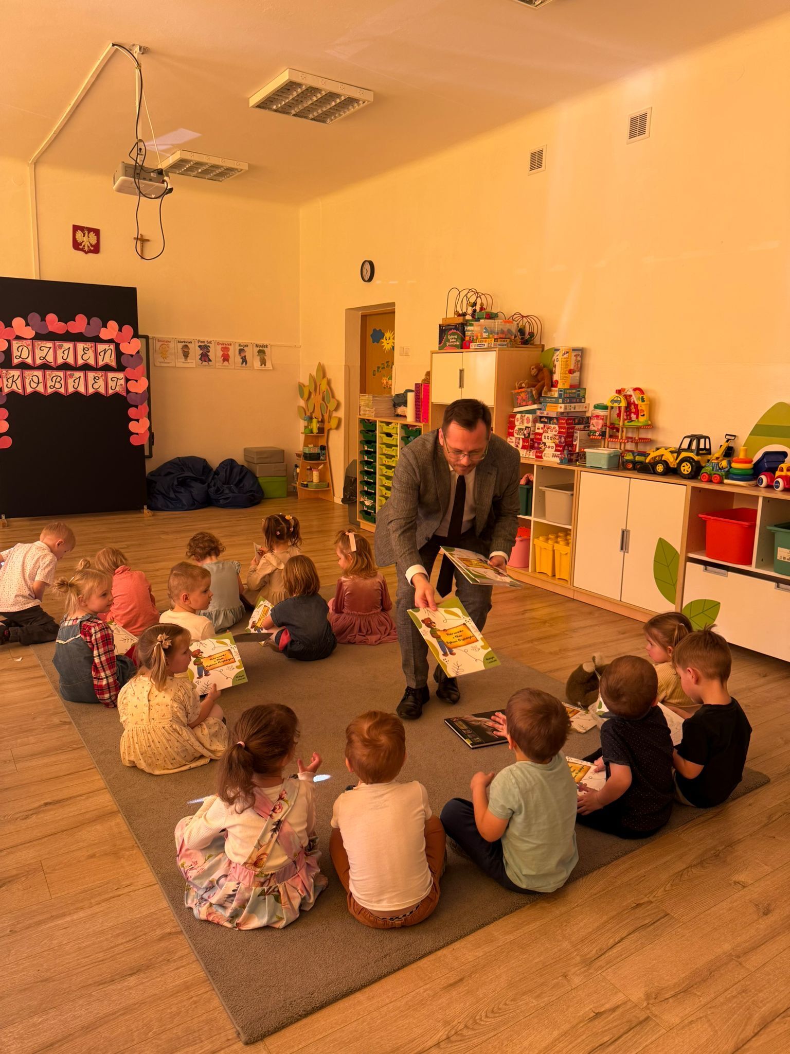 Sala przedszkolna, w której grupa małych dzieci siedzi w kręgu na dużym szarym dywanie, a dorosły mężczyzna w garniturze rozdaje im kolorowe książki lub zeszyty.  Dzieci siedzą głównie tyłem lub bokiem do aparatu, część trzyma otwarte materiały w rękach. Po prawej stronie stoją niskie białe szafki i półki z pojemnikami oraz wieloma zabawkami, w tym pojazdami budowlanymi i grami/pudełkami. W tle widać drzwi, zegar na ścianie, rysunki lub plansze edukacyjne oraz pufy. Po lewej stronie jest duża czarna tablica/dekoracja z sercami i napisem „DZIEŃ KOBIET”. Na ścianie wisi też godło Polski. Na suficie są lampy i podwieszony projektor.