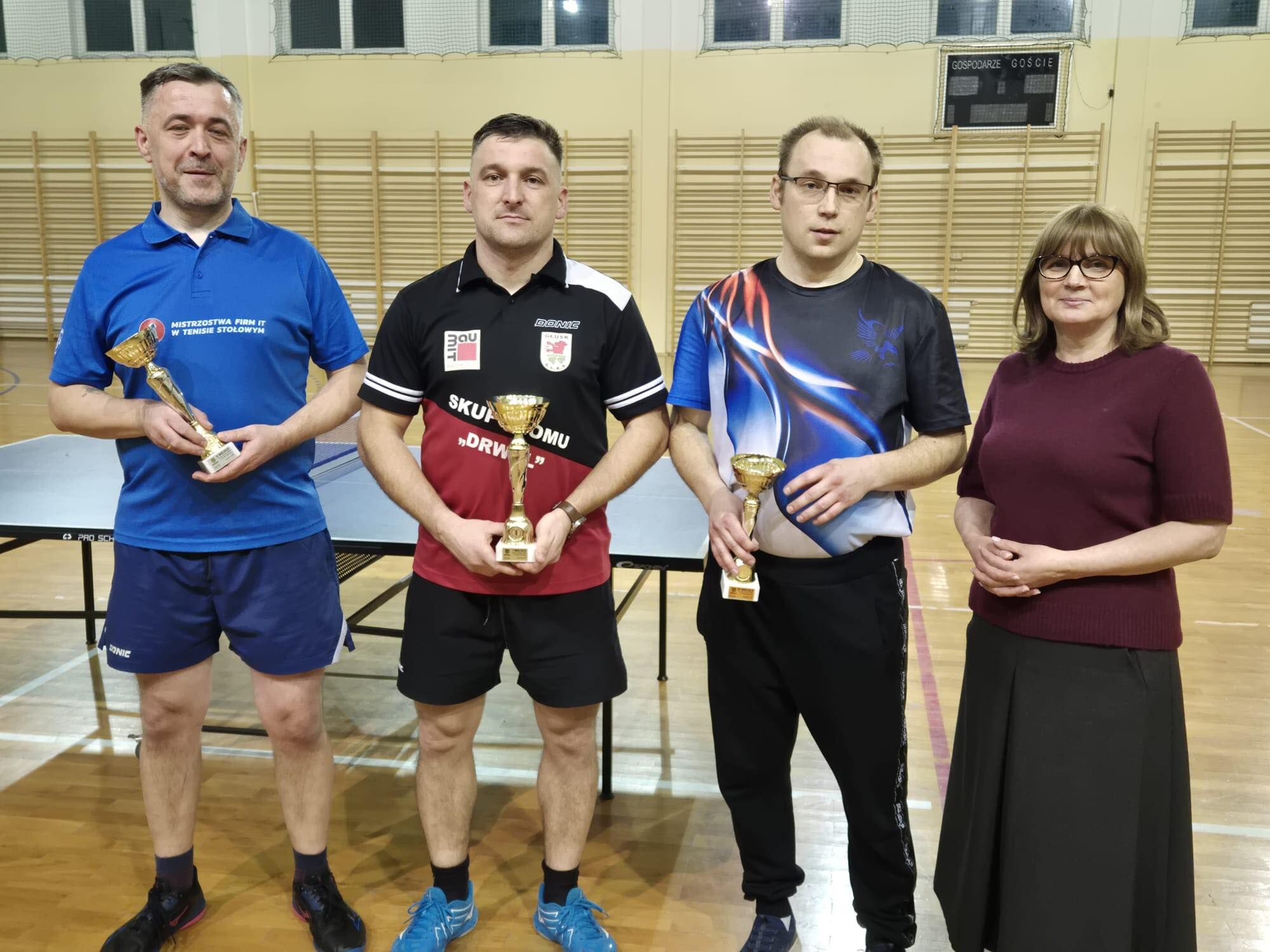 Cztery osoby stoją w sali gimnastycznej przy stołach do tenisa stołowego.  Trzech mężczyzn trzyma złote puchary: Po lewej mężczyzna w niebieskiej koszulce polo i niebieskich spodenkach sportowych; na koszulce jest napis: „MISTRZOSTWA FIRM IT W TENISIE STOŁOWYM”. W środku mężczyzna w czarno-czerwonej koszulce sportowej i czarnych spodenkach. Po prawej mężczyzna w koszulce sportowej z niebiesko-białym wzorem i czarnych spodniach; ma okulary.  Obok nich (po prawej) stoi kobieta w okularach, w bordowym swetrze i długiej ciemnej spódnicy.  W tle widać stoły do ping-ponga i drabinki gimnastyczne na ścianie; wysoko jest tablica wyników z napisami „GOSPODARZE” i „GOŚCIE”.