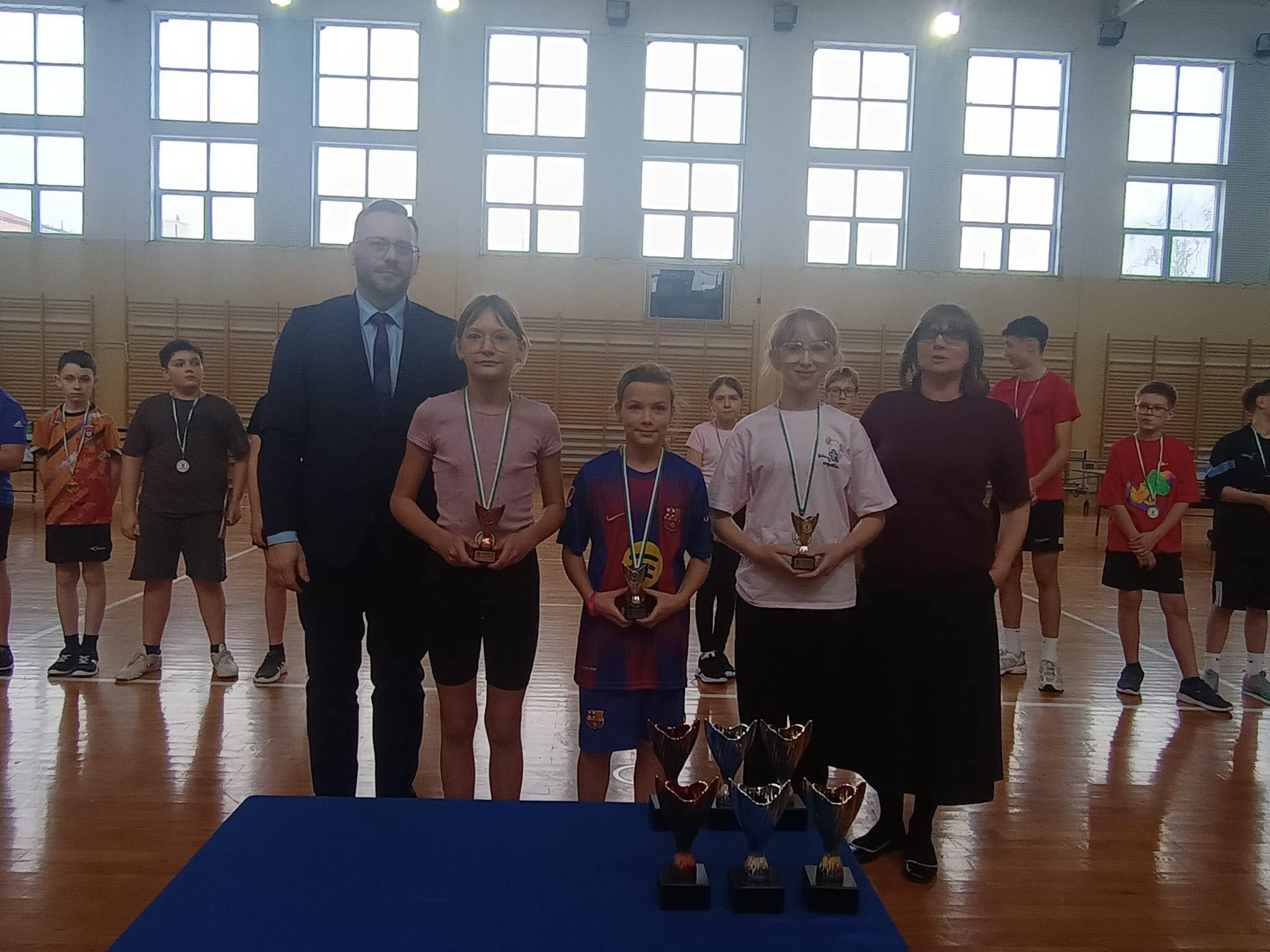 Na zdjęciu w sali gimnastycznej widać wręczenie nagród.  Na pierwszym planie stoi pięć osób w jednym rzędzie: Mężczyzna w ciemnym garniturze, białej koszuli i krawacie, w okularach. Dziewczynka w różowej koszulce i ciemnych spodenkach, ma medal na szyi i trzyma mały puchar. Dziecko w stroju piłkarskim Barcelony (granatowo-czerwony), ma medal i trzyma mały puchar. Dziewczynka w białej koszulce i ciemnych spodenkach, w okularach, ma medal i trzyma mały puchar. Kobieta w okularach, w bordowym swetrze i długiej ciemnej spódnicy.  Na stole z przodu (niebieski blat) stoi kilka dużych pucharów.  W tle widać innych uczestników z medalami, drabinki na ścianie oraz duże okna.