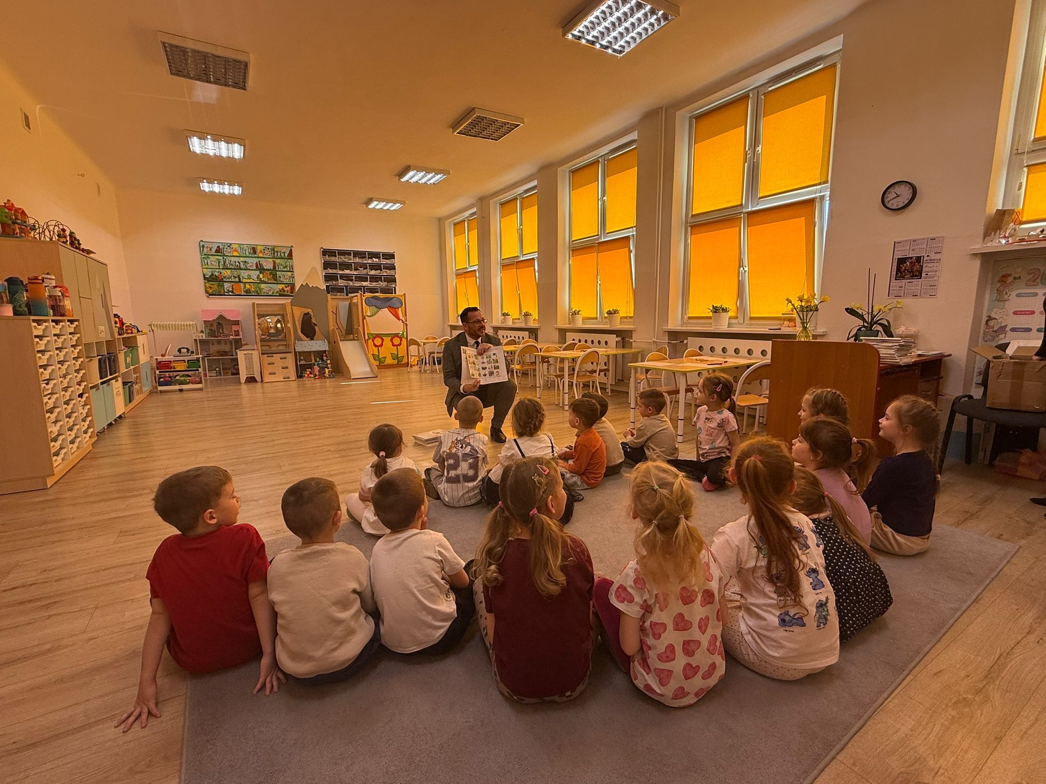 Sala przedszkolna z grupą małych dzieci siedzących w półkolu na dużym szarym dywanie. W środku, naprzeciw dzieci, klęczy dorosły mężczyzna w garniturze i trzyma otwartą książkę z obrazkami, jak podczas czytania lub prezentacji. Dzieci są zwrócone w jego stronę, większość siedzi na podłodze; część ma związane włosy w kucyki, ubrania są codzienne, w jasnych i kolorowych barwach.  Po prawej stronie znajdują się wysokie okna z intensywnie pomarańczowymi roletami, a pod oknami stoją rzędy małych stolików i krzeseł. Na ścianie po prawej wisi zegar. W głębi i po lewej stronie widać regały, półki i szafki z zabawkami oraz materiałami plastycznymi, a także kolorowe dekoracje i tablice z pracami lub ilustracjami. Podłoga jest drewniana, a na suficie są prostokątne lampy.