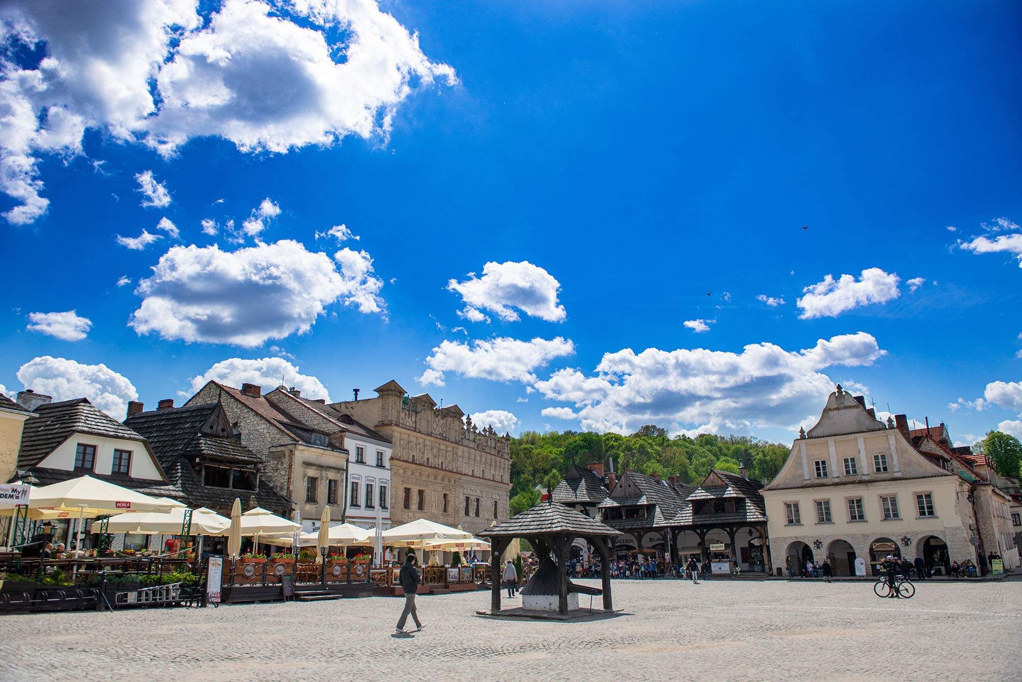 Malowniczy rynek w Kazimierzu Dolnym z zabytkowymi budynkami, brukowanym placem i altaną pośrodku; ludzie spacerują i jeżdżą na rowerze pod błękitnym niebem pełnym białych chmur.